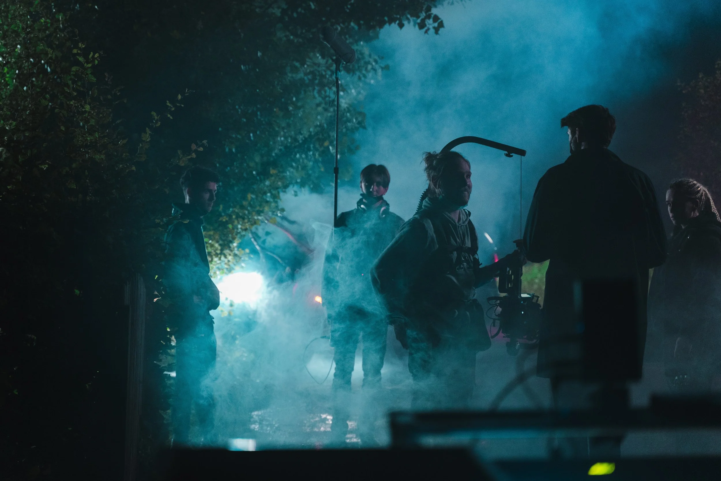 Film crew working outdoors at night with smoky atmosphere, surrounded by trees