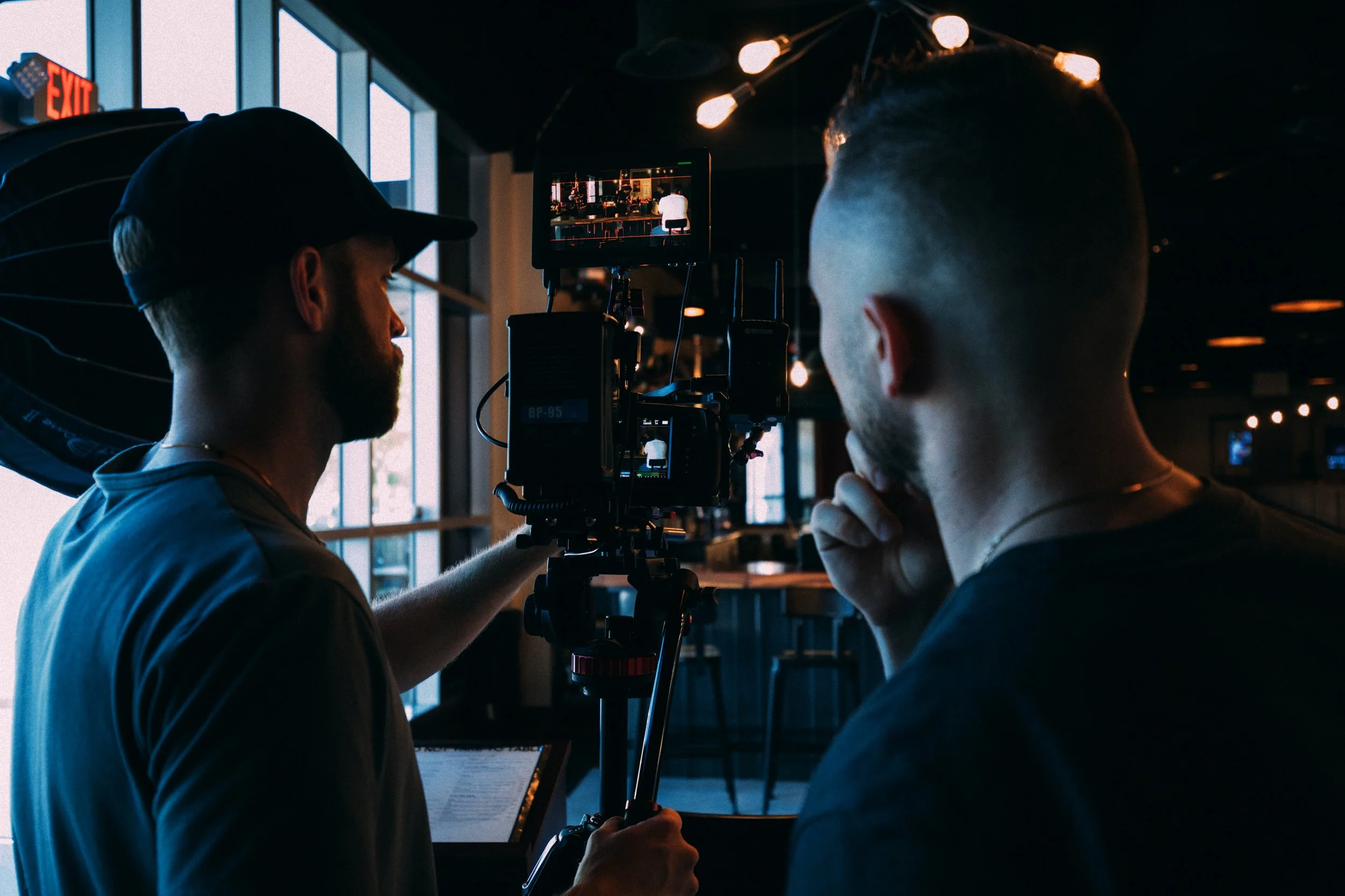 Two men working on a film or photography set inside a dimly lit restaurant or cafe, with large windows and a decorative chandelier overhead.