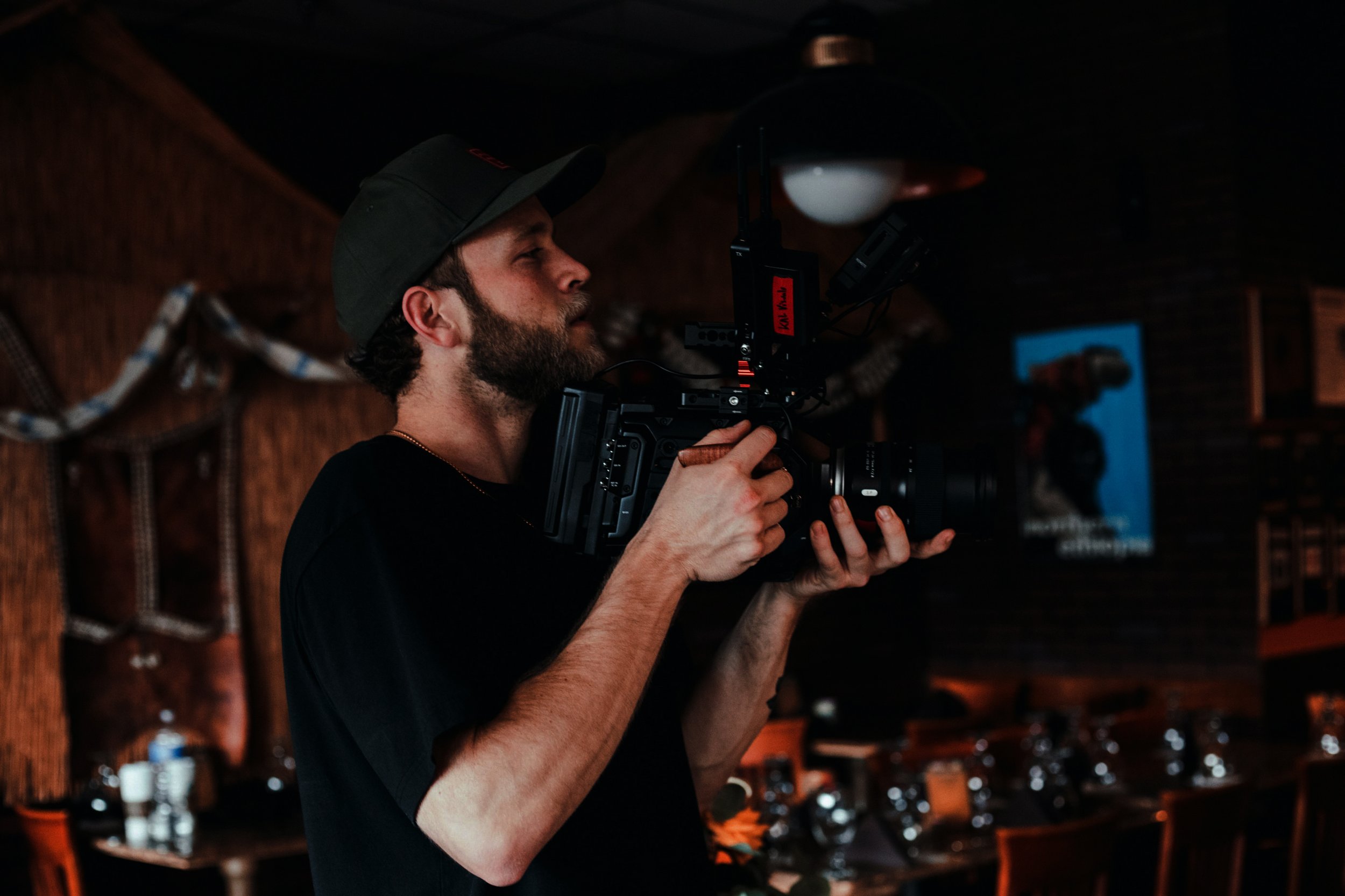 A man with a beard wearing a black shirt and a cap operating a professional video camera in a dimly lit indoor space.