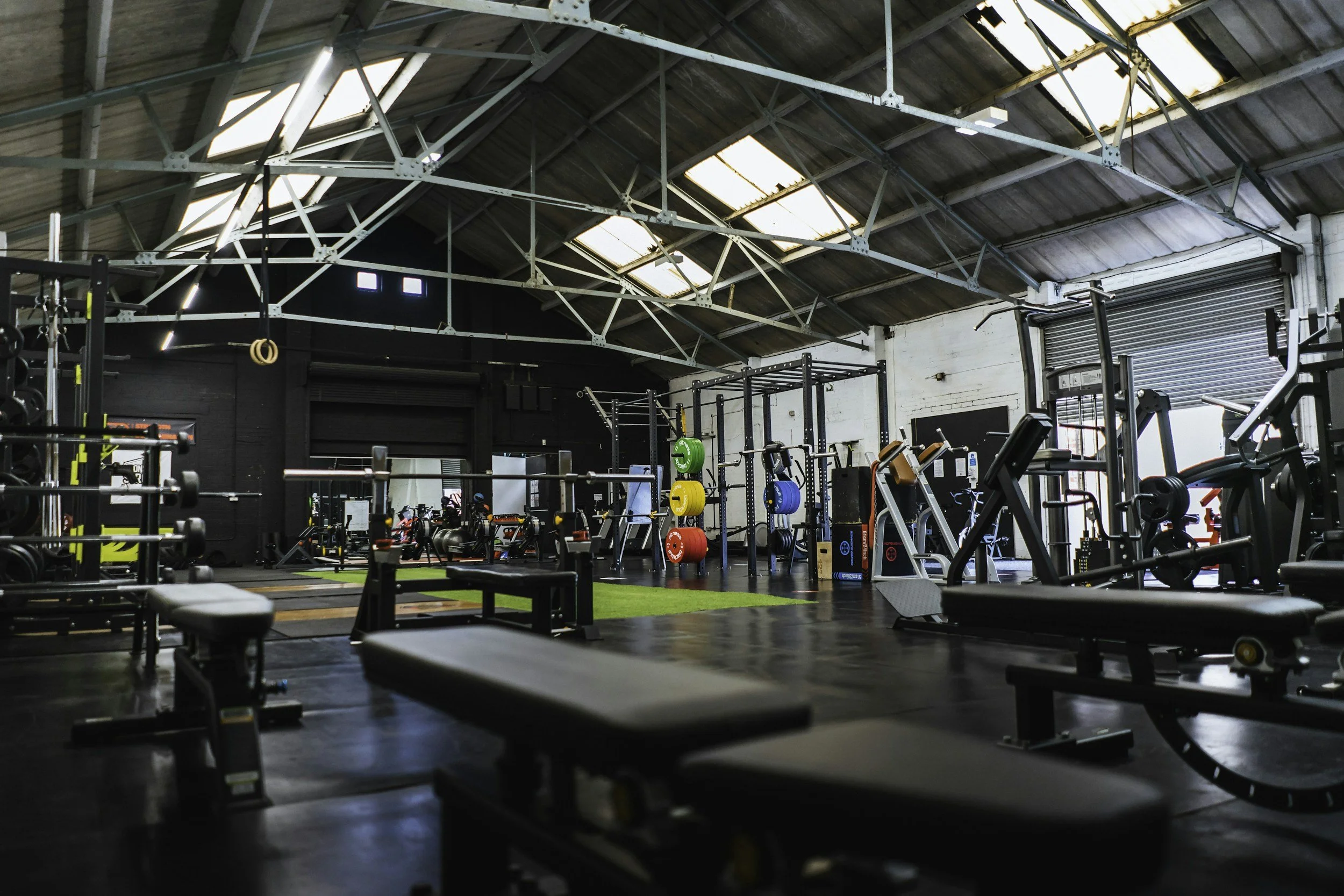 Empty gym with various workout equipment, benches, weights, and exercise machines inside a spacious industrial-style building with high ceilings and skylights.