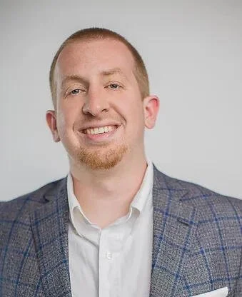 A young man with short red hair, a light beard, smiling, wearing a checkered blazer and white shirt, against a plain light gray background.