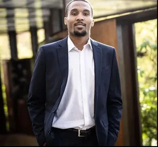 Man in a navy suit and white shirt standing outdoors in front of a wooden structure and trees.