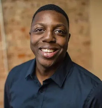 A smiling man with dark skin, short hair, wearing a dark blue collared shirt, standing in front of a brick wall.