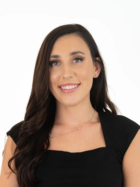 A woman with long dark brown hair wearing a black top and a delicate necklace, smiling against a plain white background.