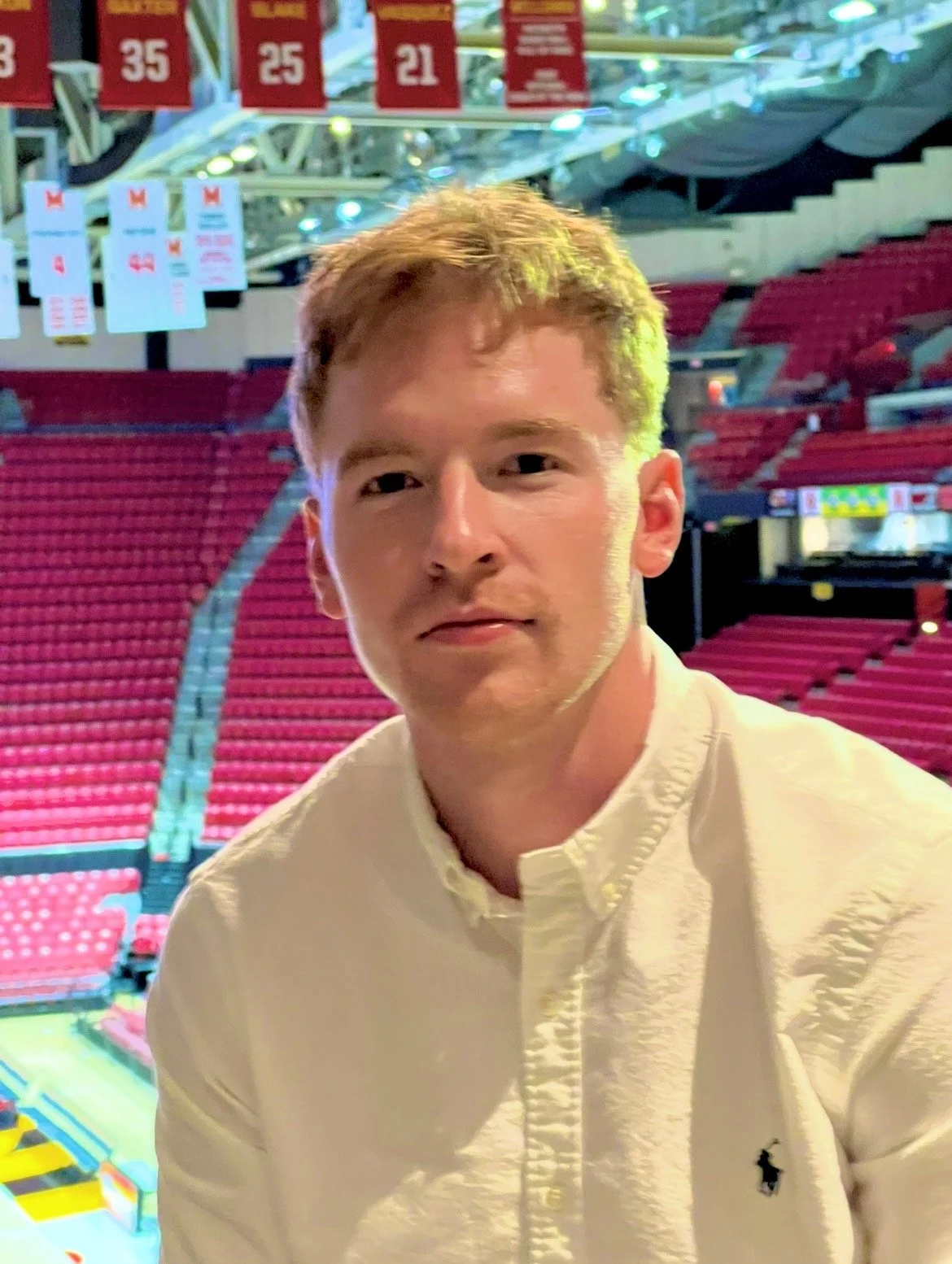 A young man with light brown hair wearing a cream-colored shirt sitting inside a sports arena with red seats, banners, and a basketball court in the background.