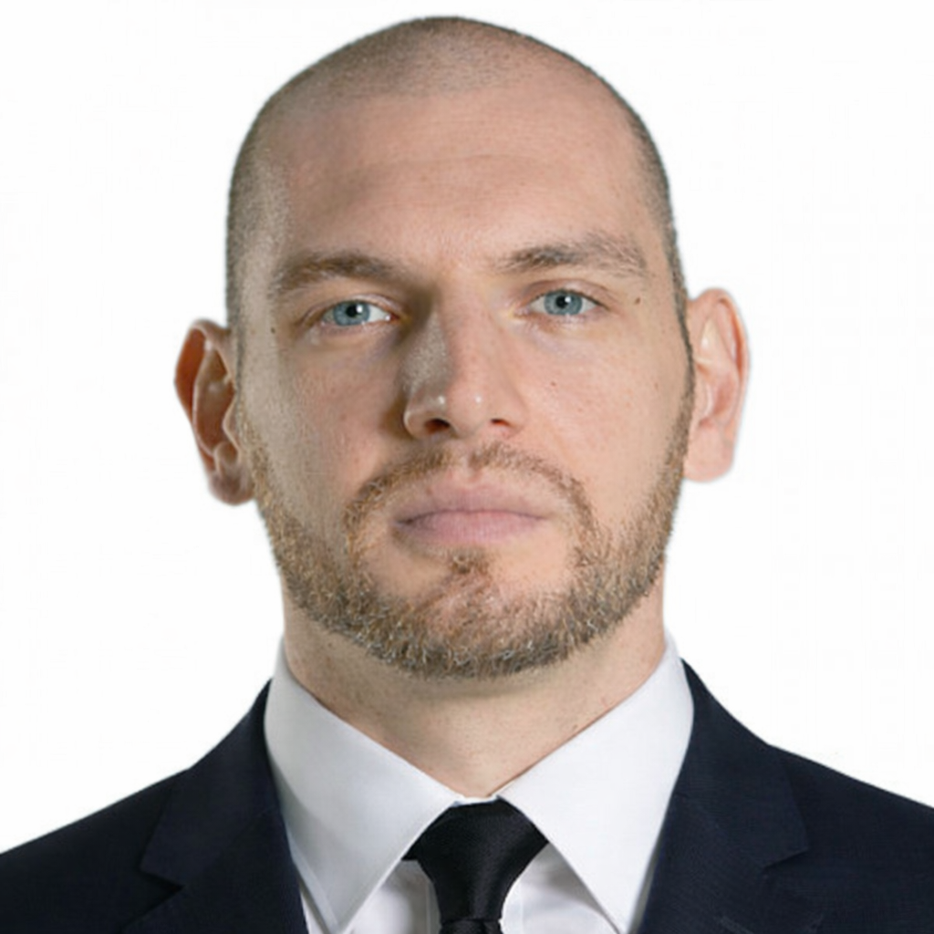 Headshot of a man with a serious expression, blue eyes, and a short beard, wearing a black suit, white shirt, and black tie, against a plain white background.