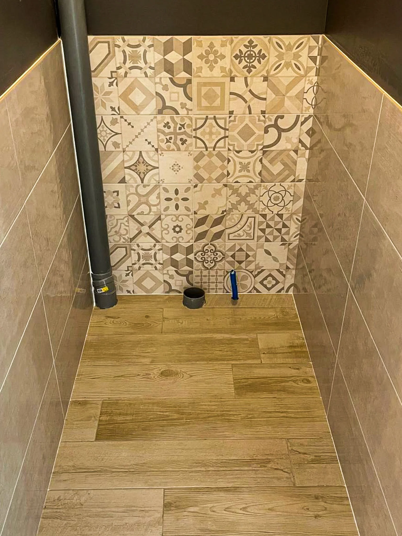 Intérieur d'une douche inachevée avec carrelage décoratif, tuyau d'évacuation et tuyau d'alimentation d'eau visibles.