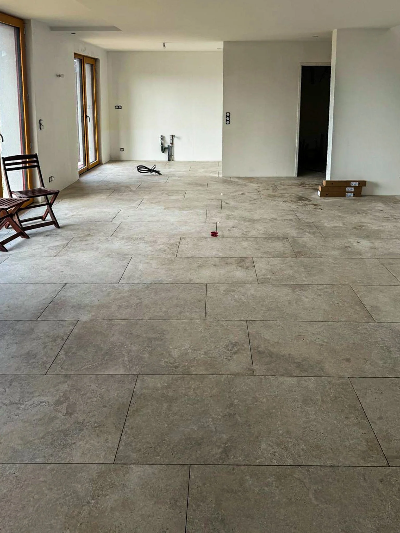 Intérieur d'un appartement en cours de rénovation avec sol en carrelage beige, murs blancs, fenêtres en bois, deux chaises en bois à gauche, et voir des Travaux en cours.