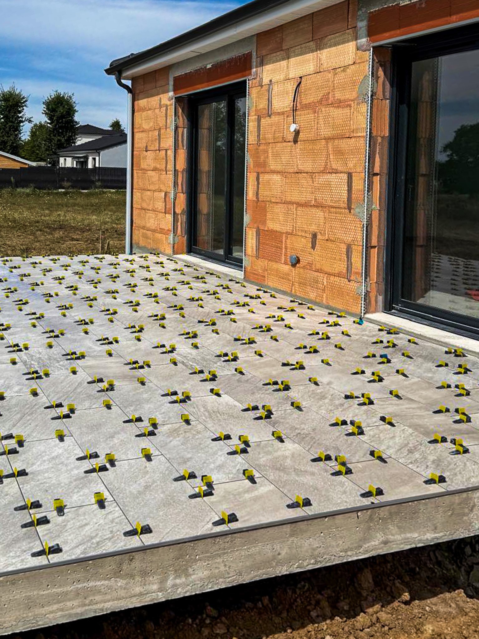 Construction d'une terrasse en béton avec des clips pour poser des lames de terrasse en bois. Maison en cours de construction avec murs en brique et fenêtres en verre