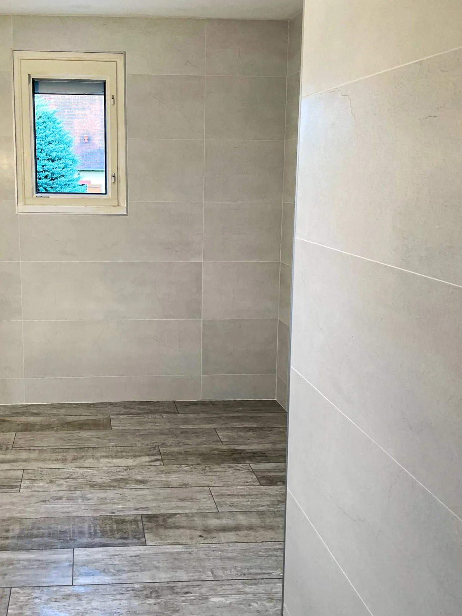 Une salle de bain ou toilettes avec un mur en carrelage beige clair, un sol en carrelage imitation bois, une fenêtre donnant sur un arbre et un bâtiment extérieur.