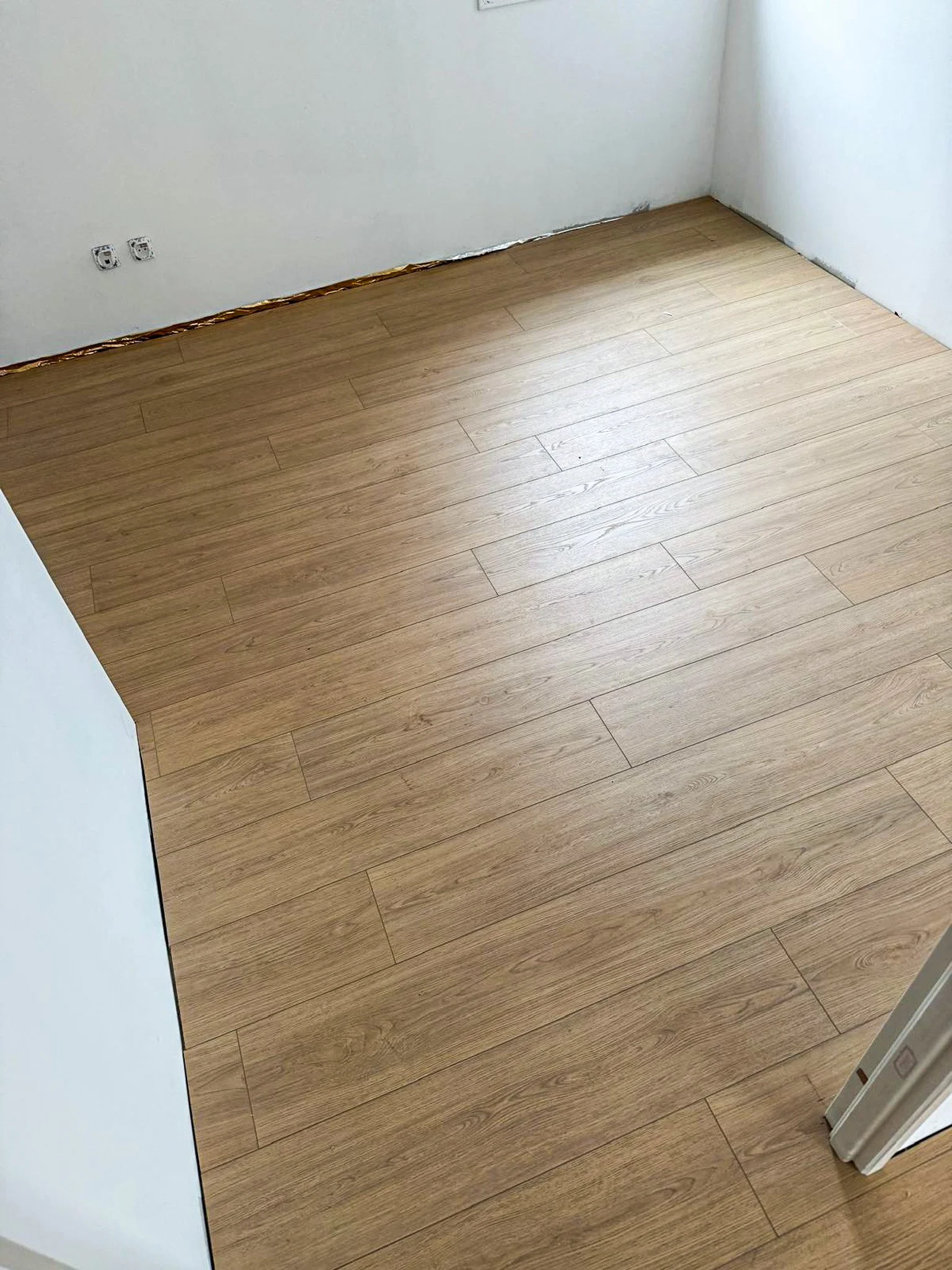 Sol en bois clair dans une pièce en construction ou rénovation, avec murs blancs et prises électriques visibles.