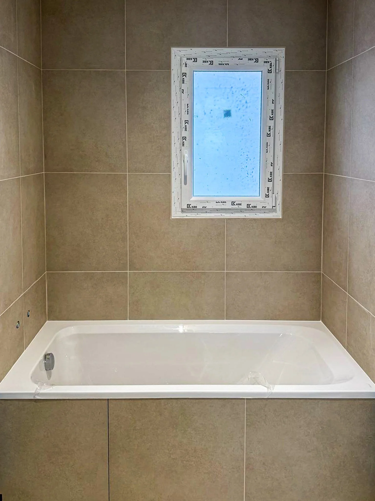 Une salle de bain avec une baignoire blanche encastrée, entourée de carrelage beige au mur. Une petite fenêtre rectangulare en haut, avec du gel ou de la condensation sur la vitre.