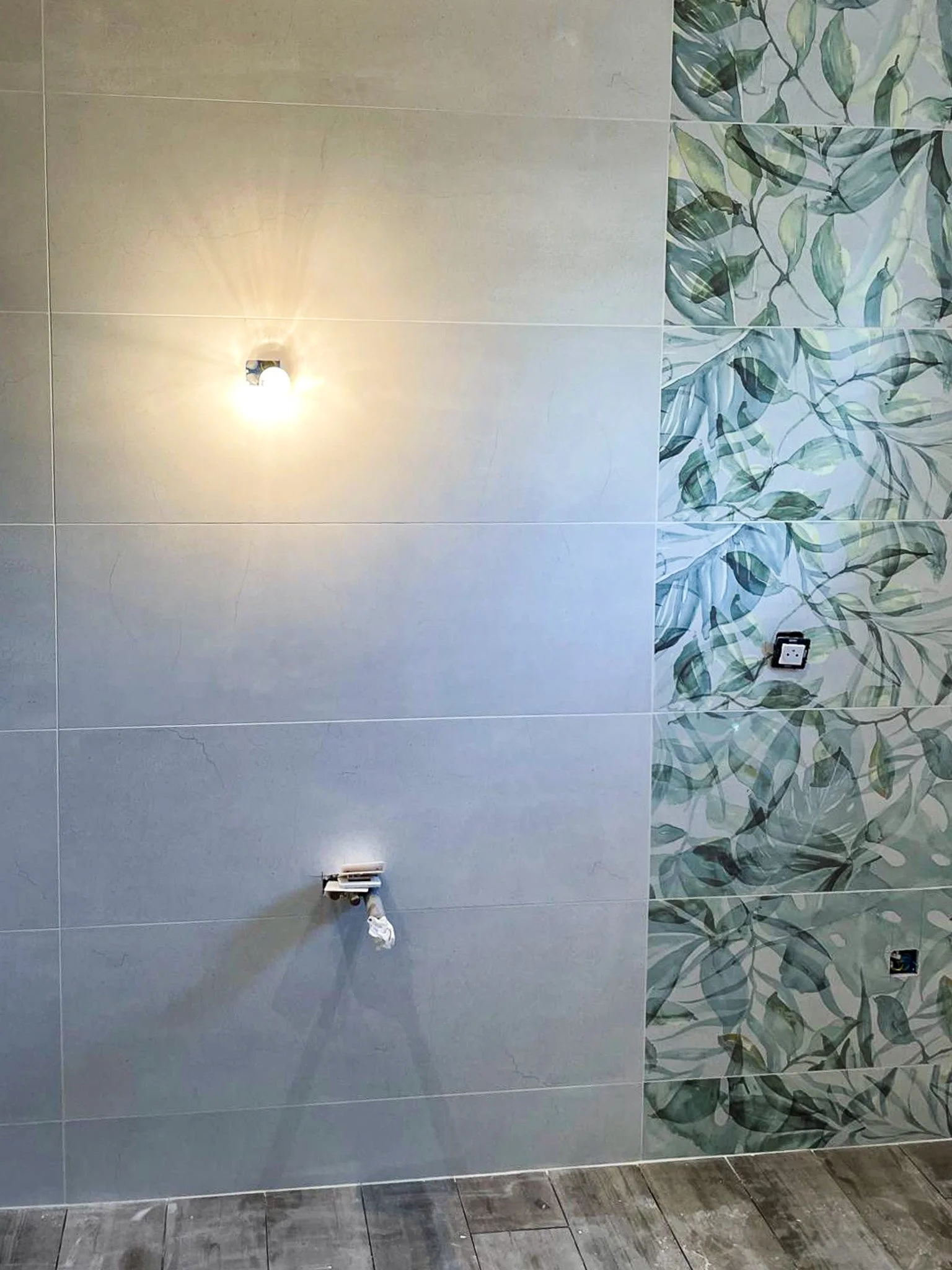 Mur en céramique gris avec une partie à motif feuilles vertes et une partie lisse, prise électrique vide, un ampoule allumée, un câble électrique suspendu.