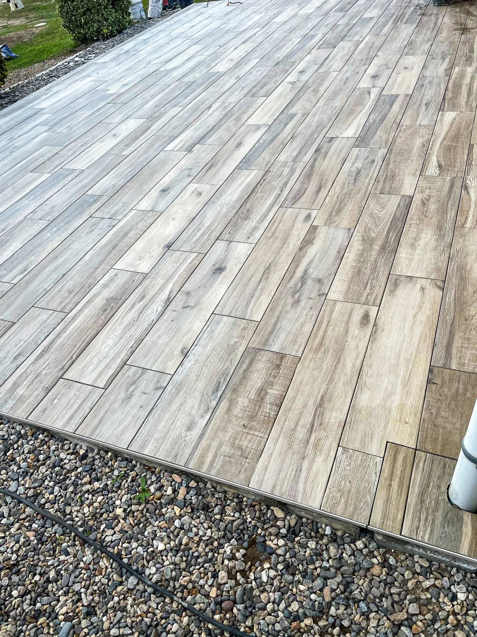 Dalles de bois claires posées en pose rectangulaire sur une terrasse ou un patio, entourées de gravier, avec un tuyau noir visible en bas.