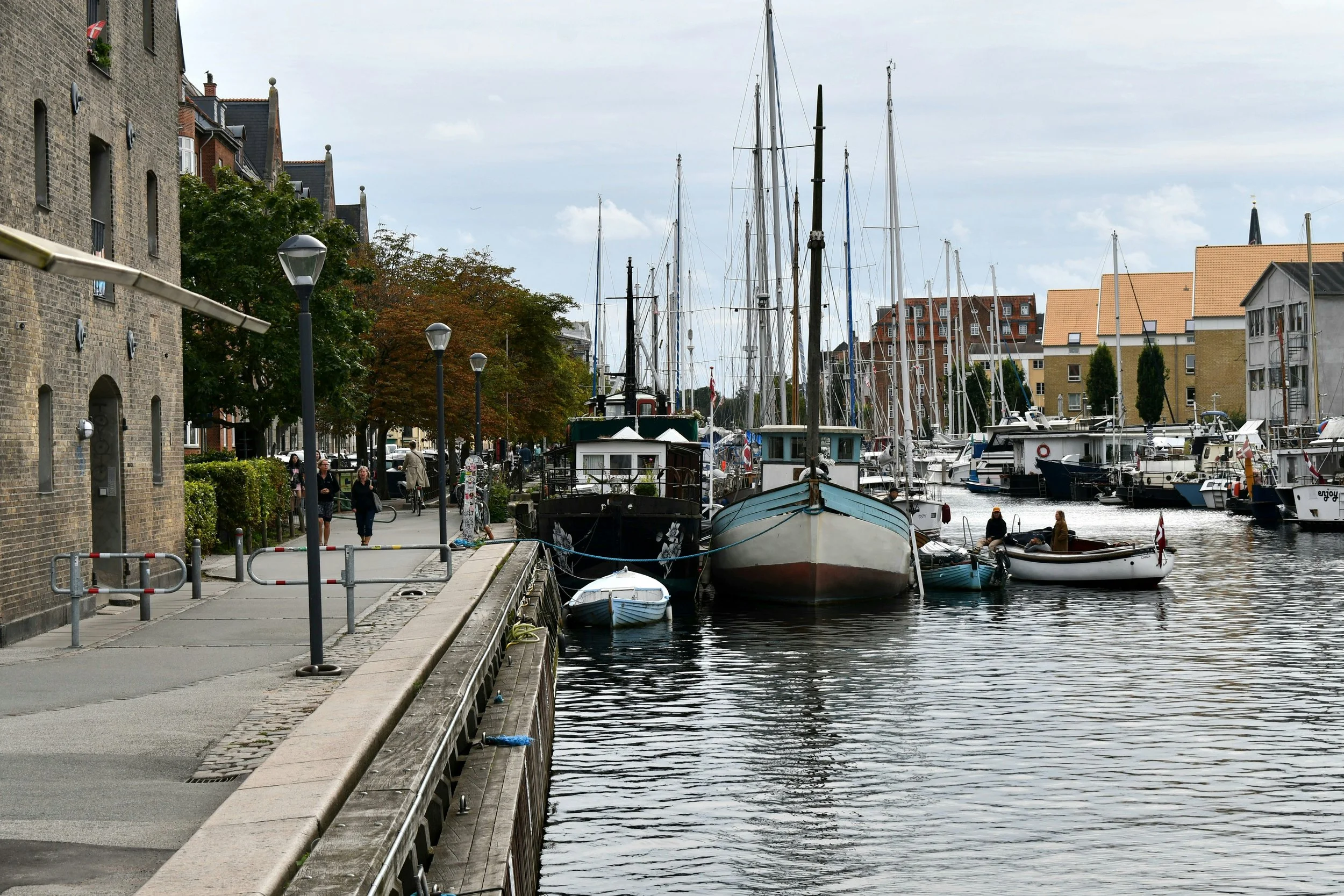 Billede af en havn med flere både, bådejere og mennesker, der går langs kajen, omgivet af bygninger og træer.