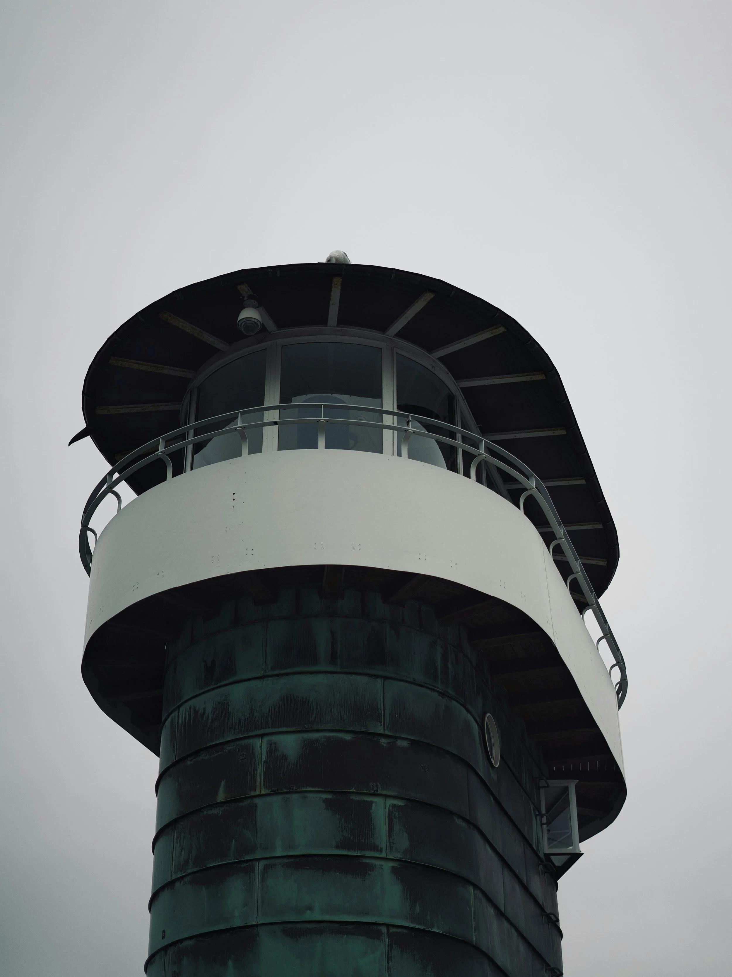 Nærbillede af et fyrtårn med en rund øvre del og et observatorium med store vinduer, maskiner og en sort kappe, under en overskyet himmel.