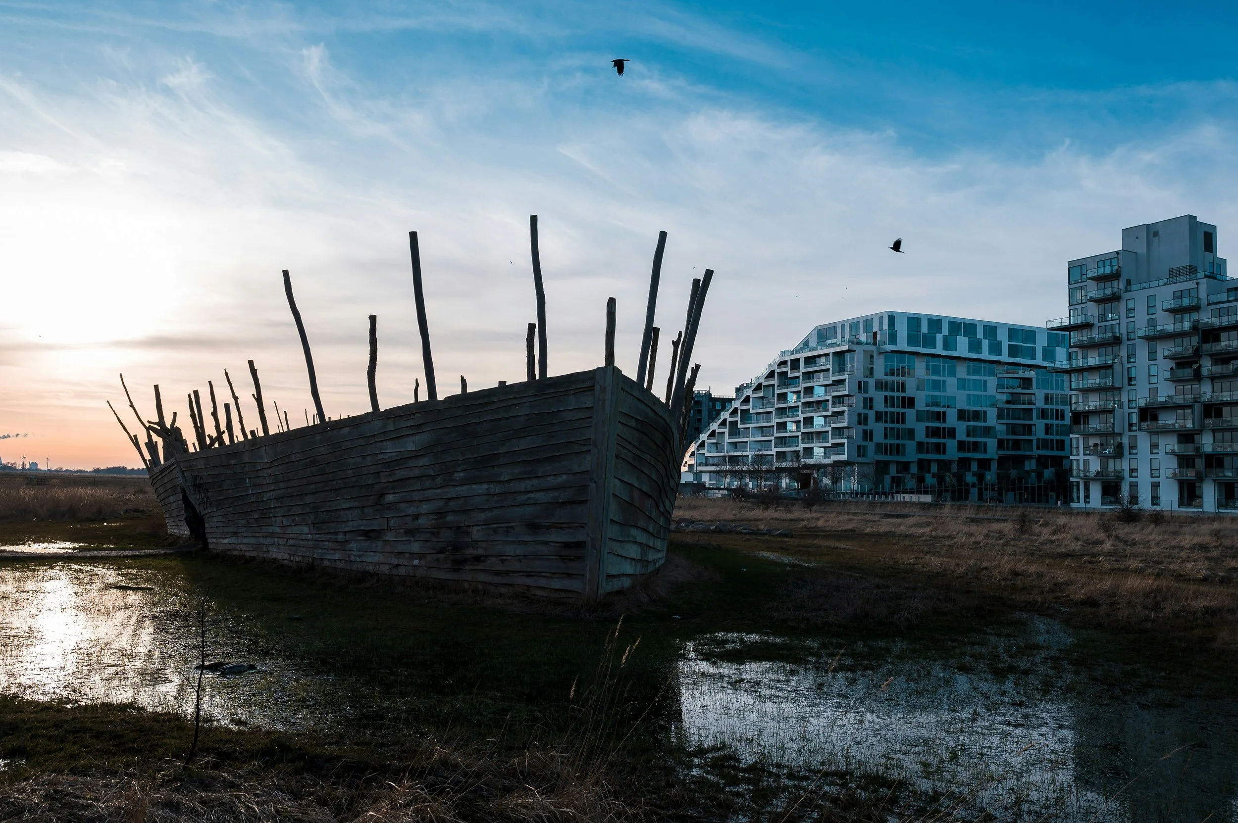 Et forladt træskib med opretstående rammer ligger på mudret jord ved vandet, med moderne boligbygninger i baggrunden under en skyet himmel. 