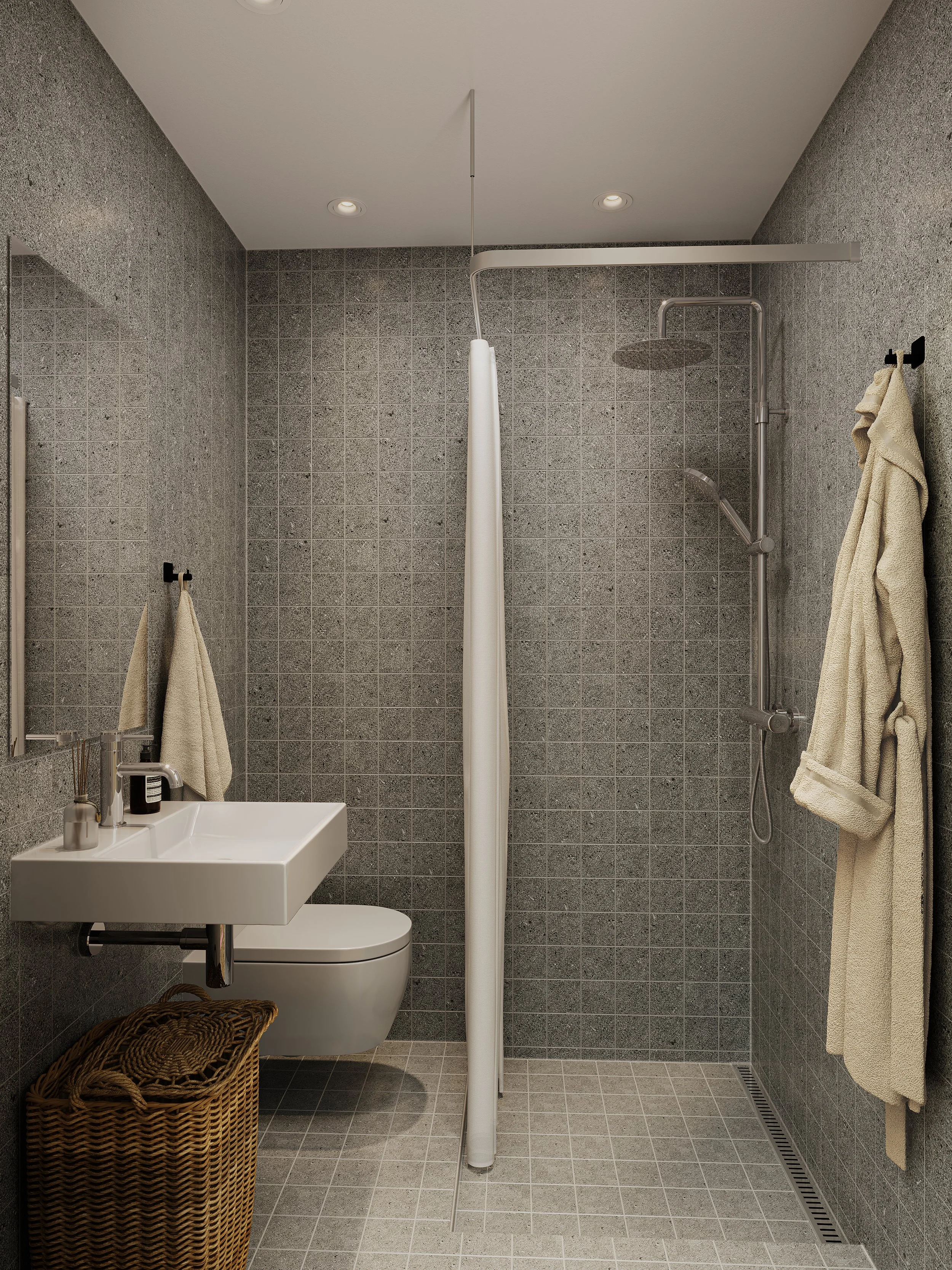 Et badeværelse med grå væg- og gulvfliser, vægmonteret toilet, smallest sink, spejl, gulvafløb, bruseområde med gardin, håndklæder på vægkroge.