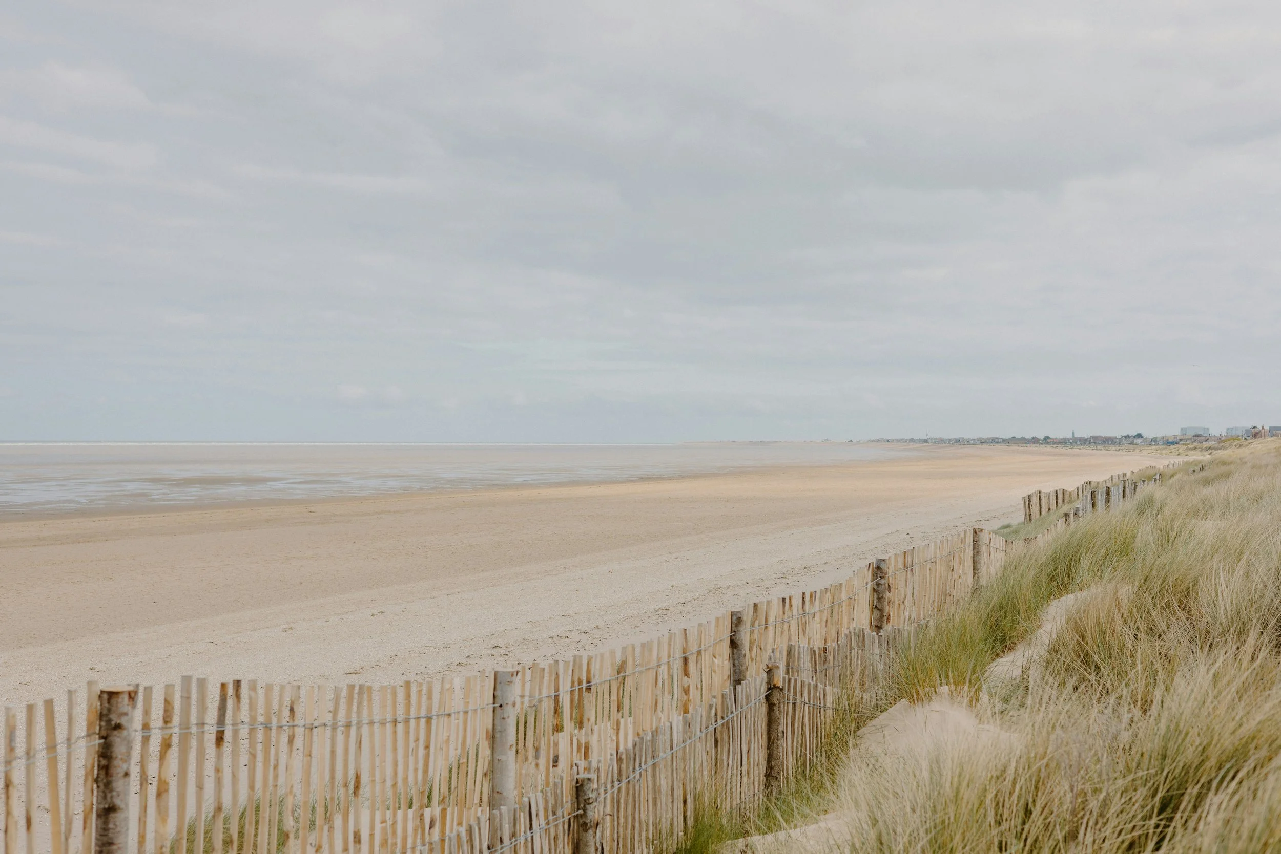 Strand med sand, høje græsser langs en trådhegn, overskyet himmel, fjerne byfacader i horisonten.