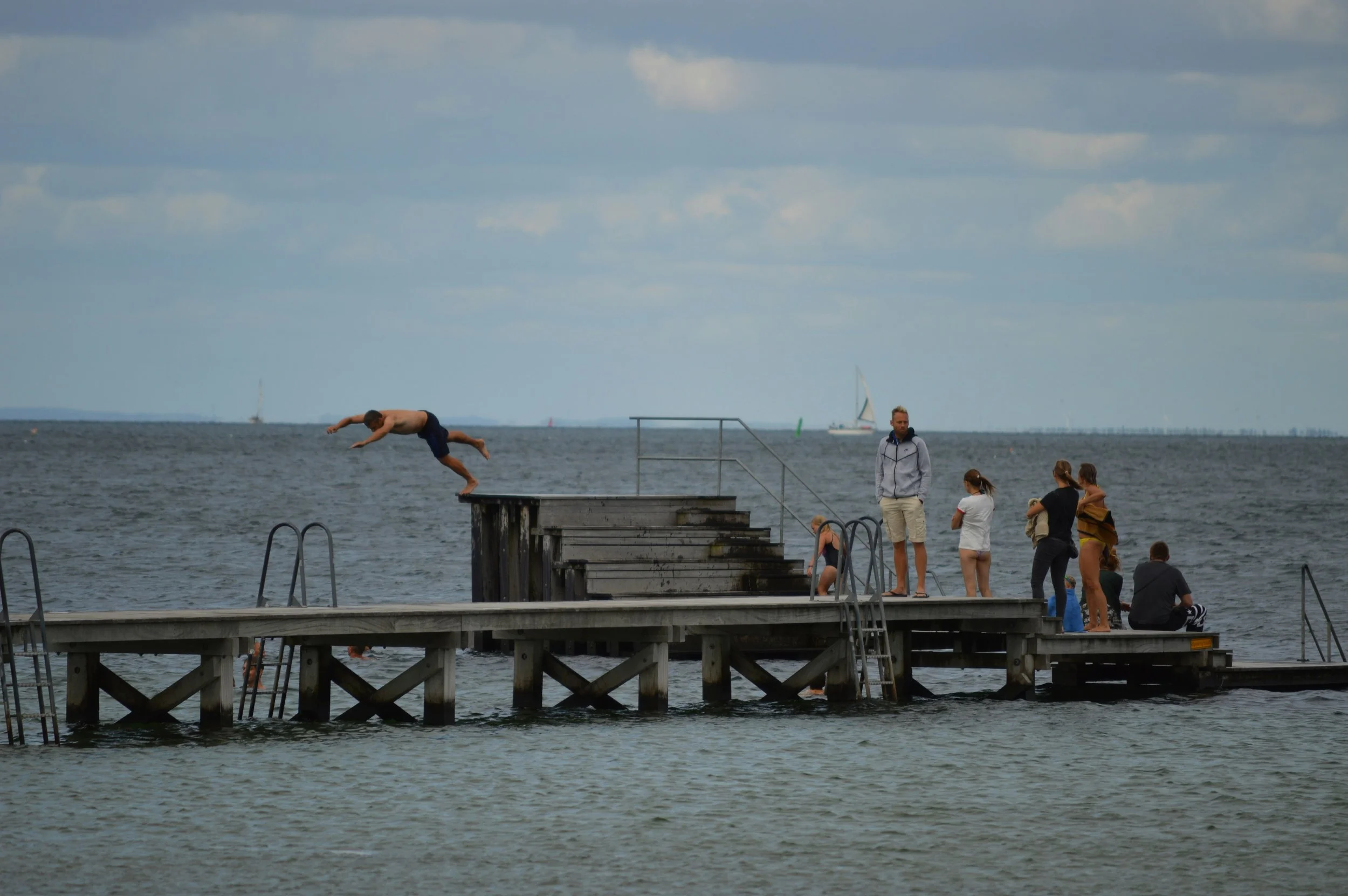 En person springer ud i vandet fra en badebro, mens flere andre mennesker står og sidder på broen ved stranden.
