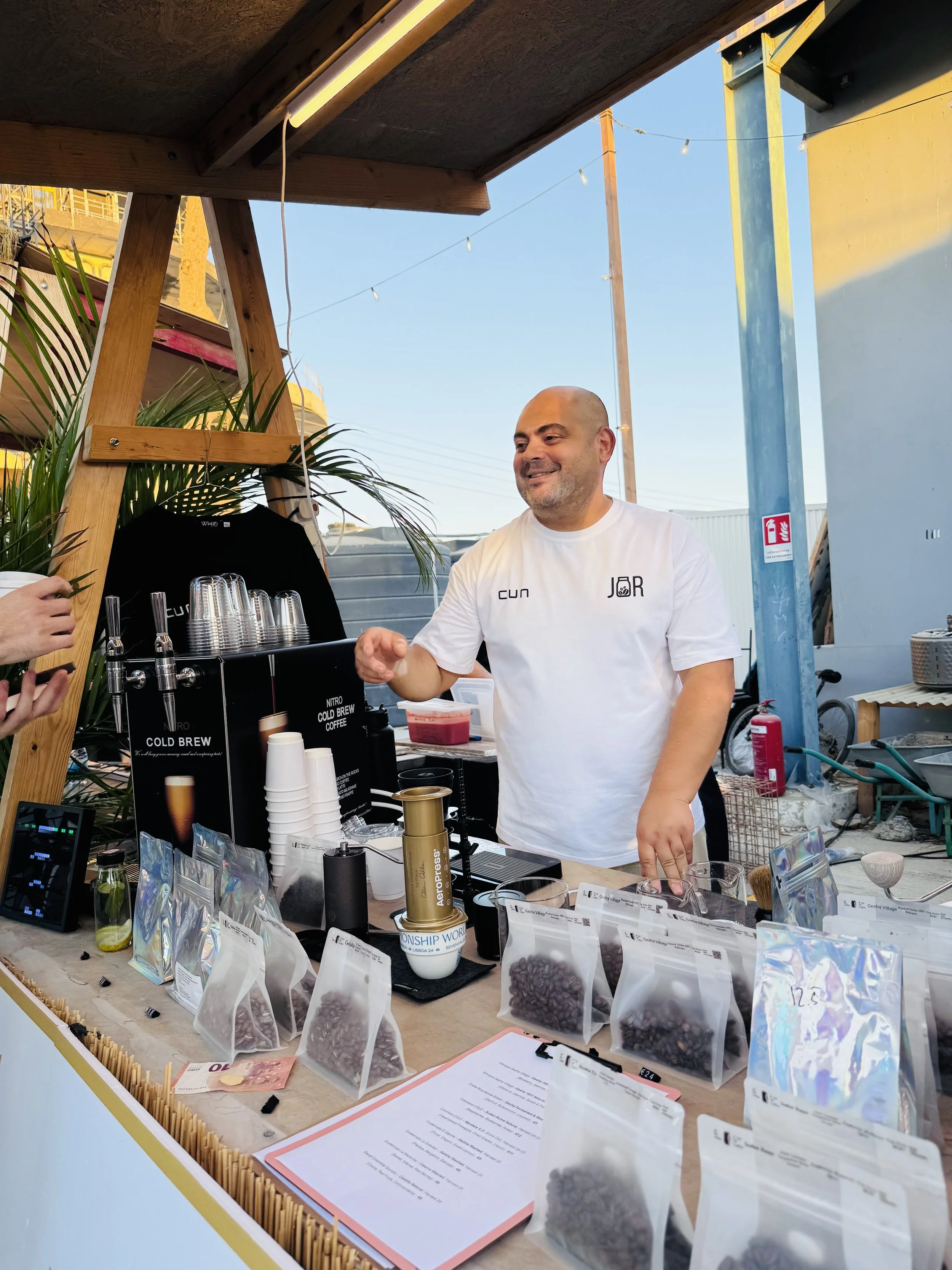Roman Vilanov, Aeropress Champion of Cyprus & Barista Hustle Coach, smiling and serving competition level coffees at Kolla.cy market in Limassol, with bags of coffee beans, a menu, and brewing equipment displayed.