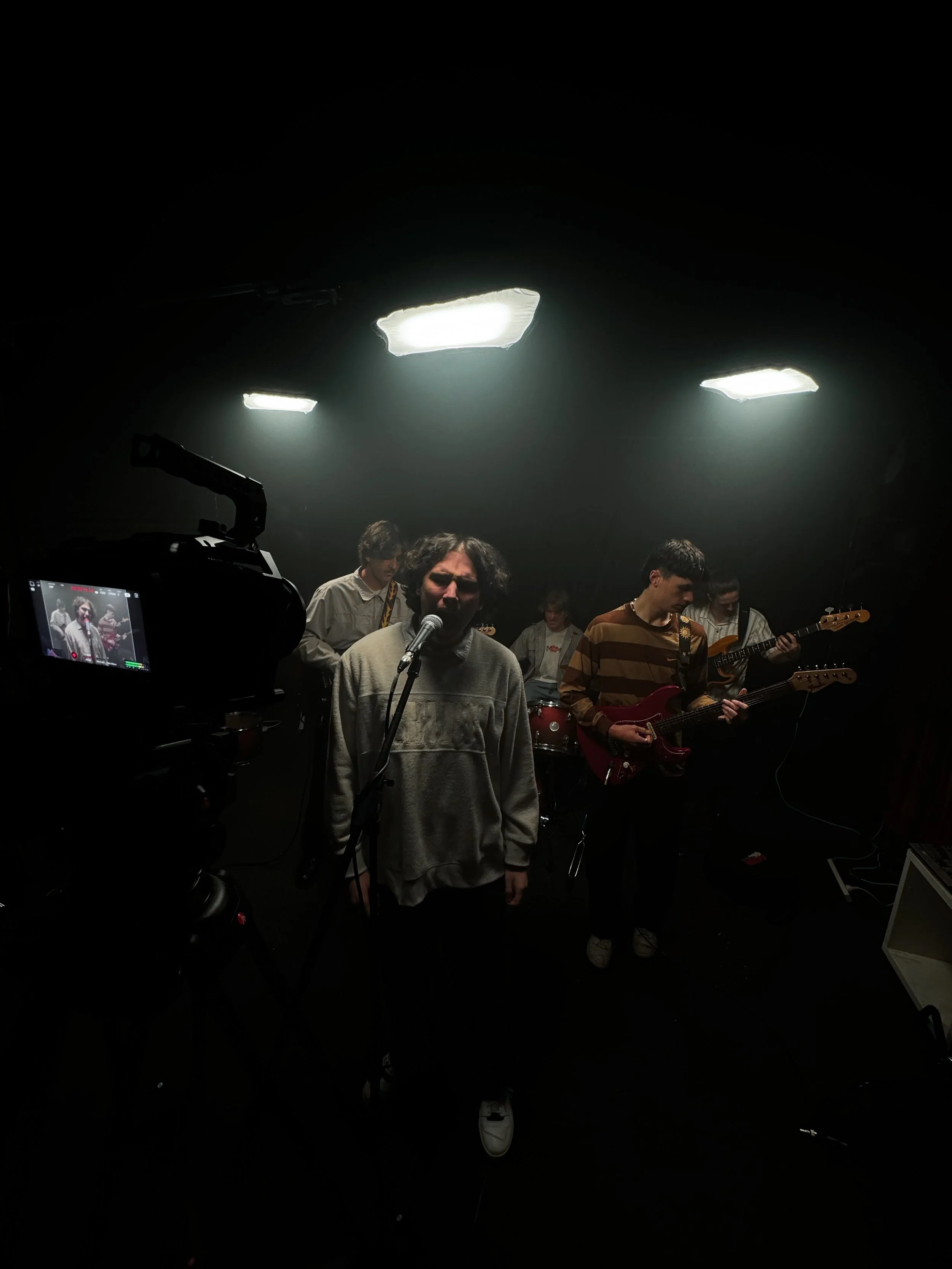 Band performing in a dark studio with bright overhead lights, including a vocalist at the microphone and guitarists in the background.