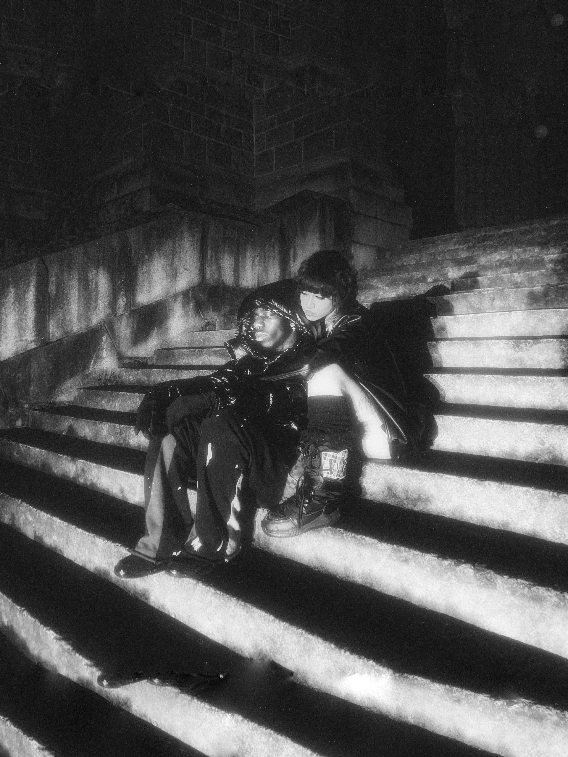 Two young people sitting on city steps at night, one resting head on the other's shoulder. Both dressed in dark clothing, with one wearing a shiny jacket and the other in sneakers. Black and white photo with shadows cast on the steps.