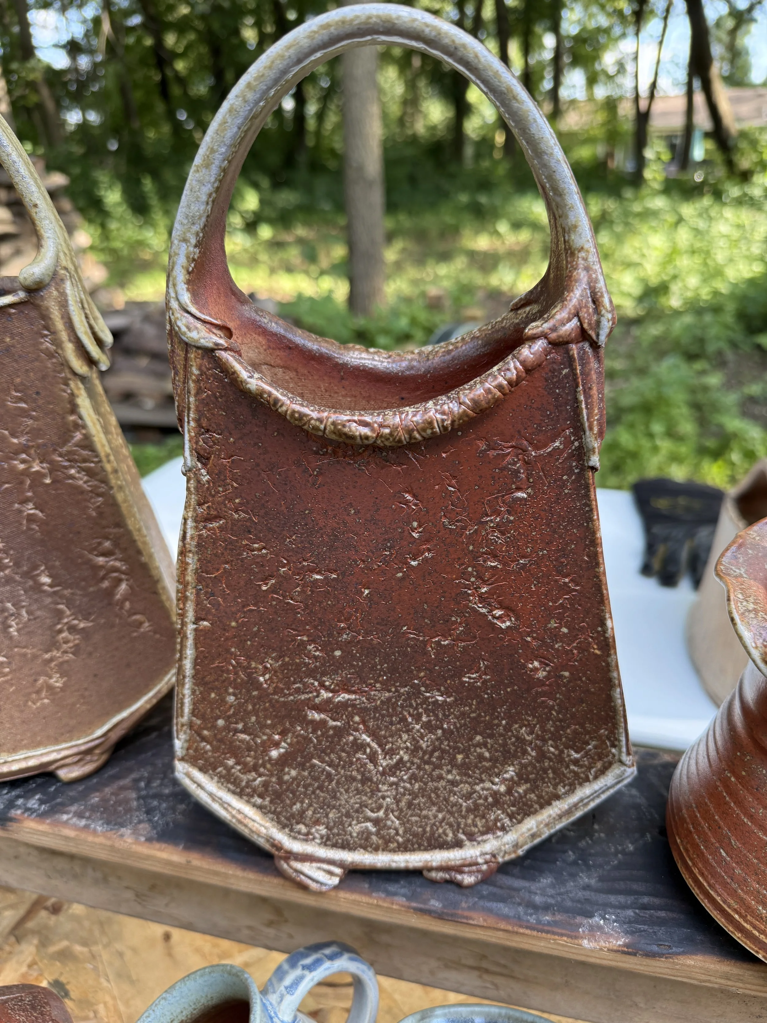 Close-up of a small, rectangular ceramic handbag-shaped planter with a textured brown glaze, set outdoors with trees and other pottery in the background.