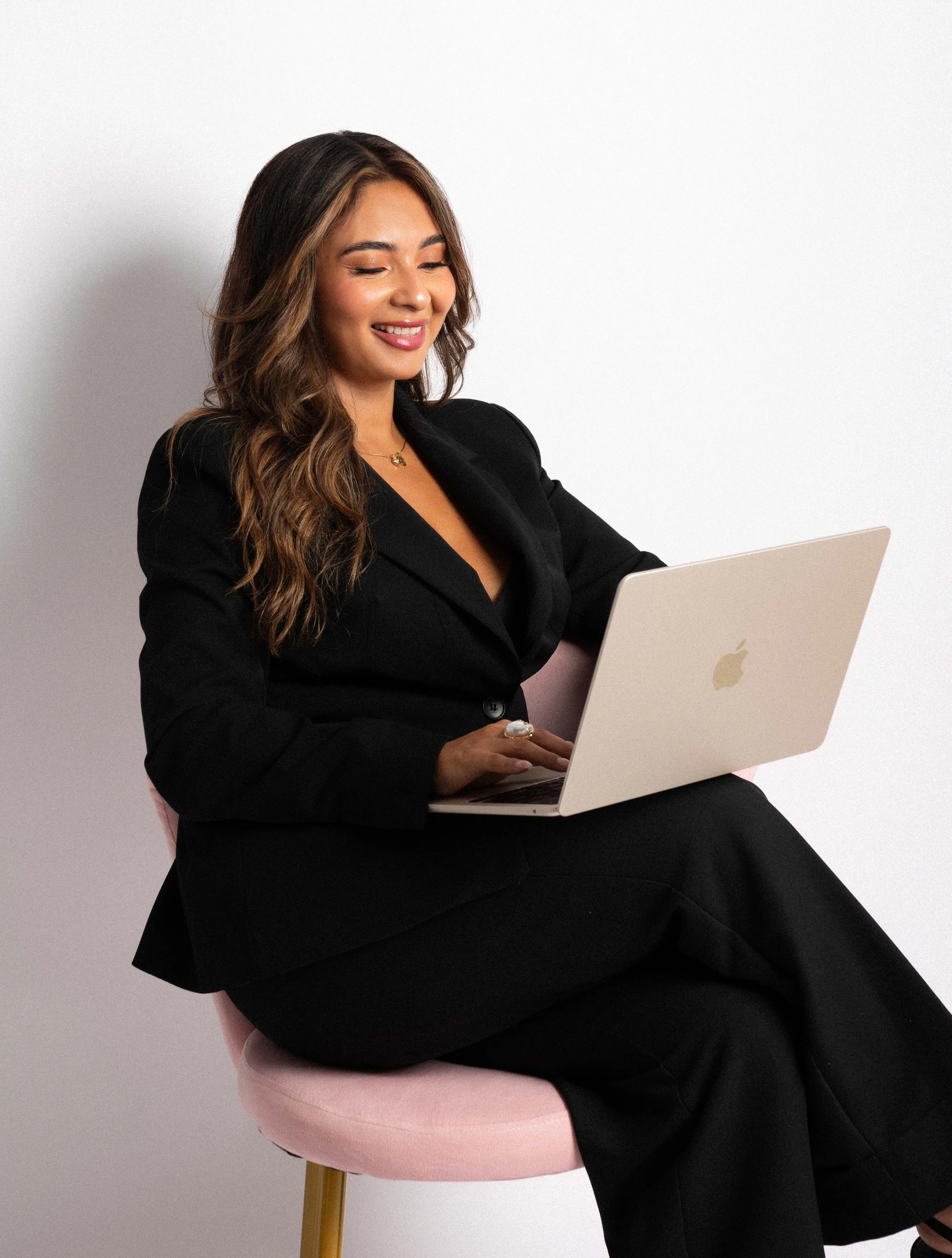 A woman with long wavy hair wearing a black suit, sitting on a pink chair, working on a silver MacBook, smiling against a plain white background.