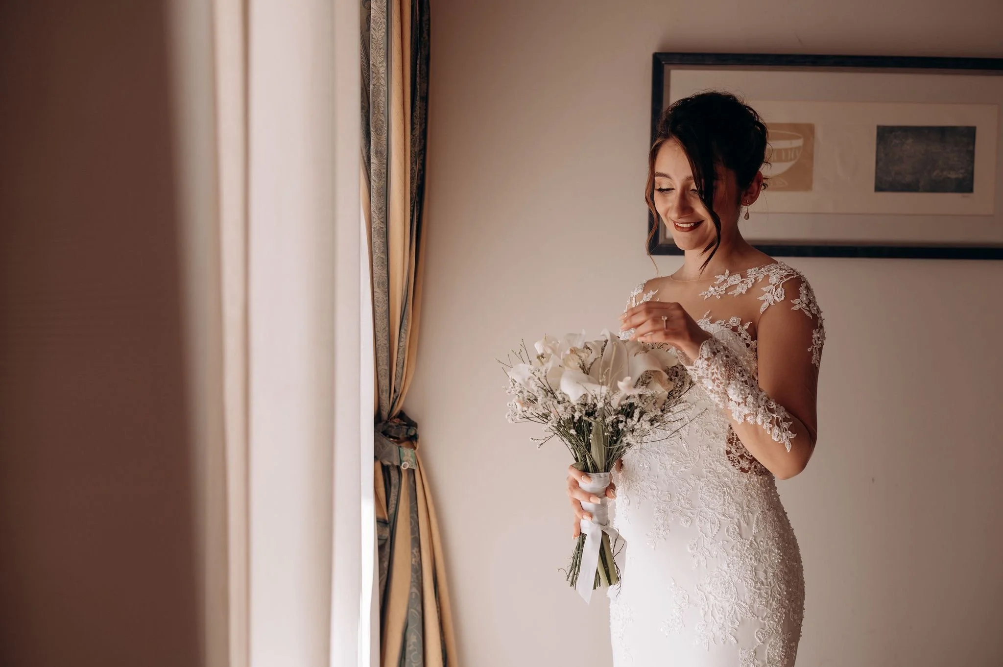 A bride in a lace wedding dress holding a bouquet of white flowers, smiling while standing near curtains and a framed picture on the wall.