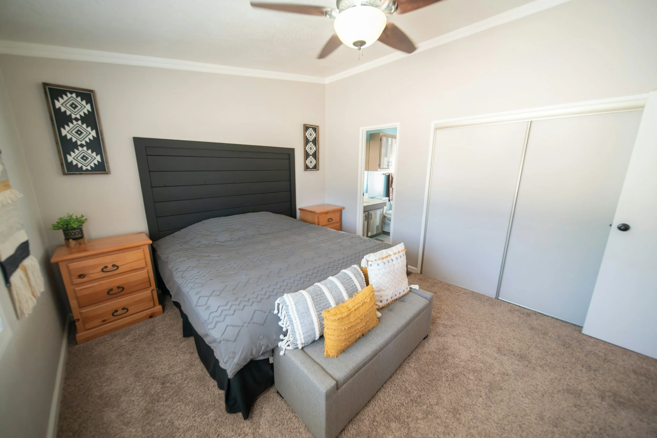 A bedroom featuring a bed with a black headboard, nightstands on each side, a ceiling fan, a decorative wall hanging, a small bench with pillows, a closet with sliding doors, and a doorway to another room.