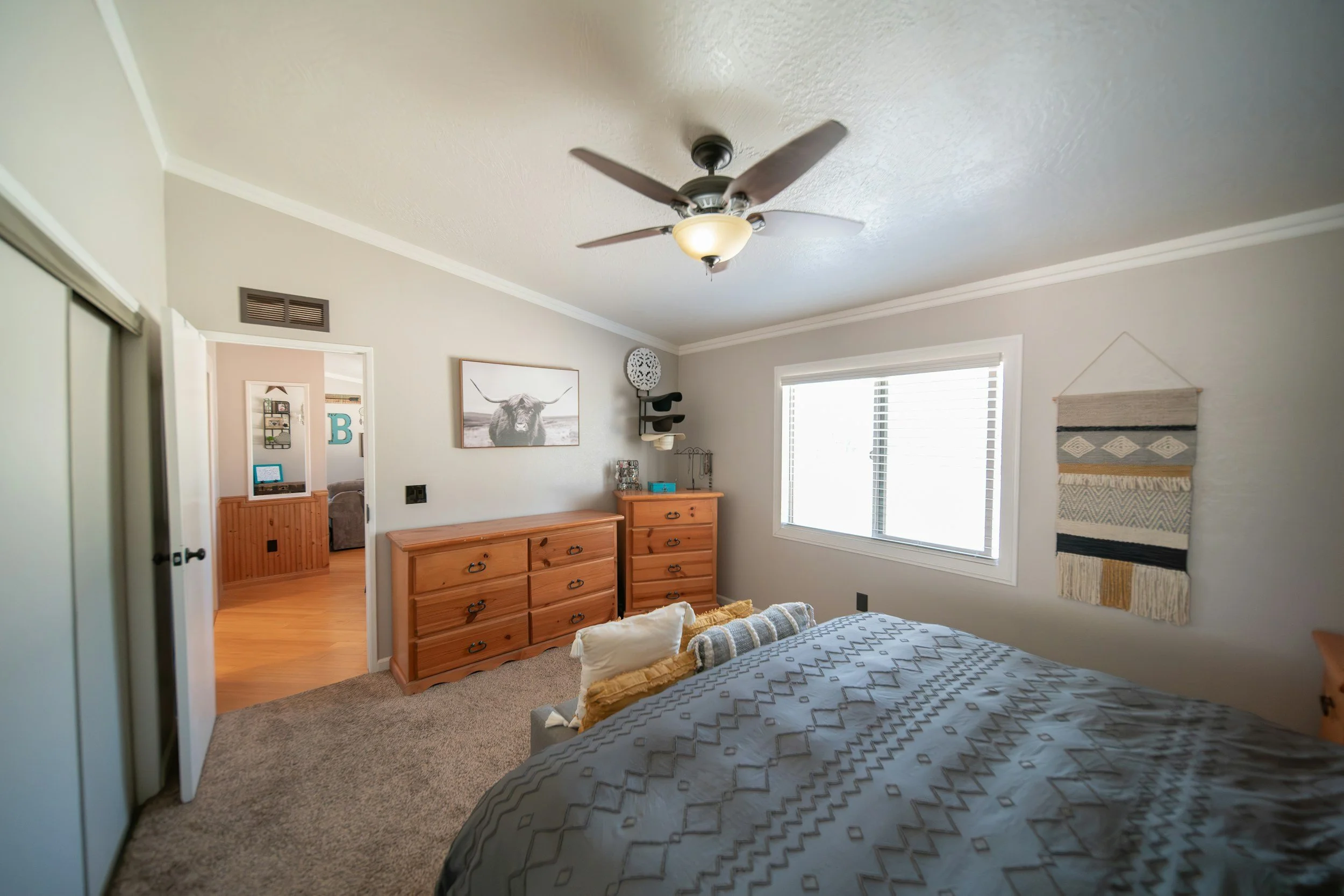 A bedroom with a bed, window, wooden dresser, wall art, ceiling fan, and a closet.