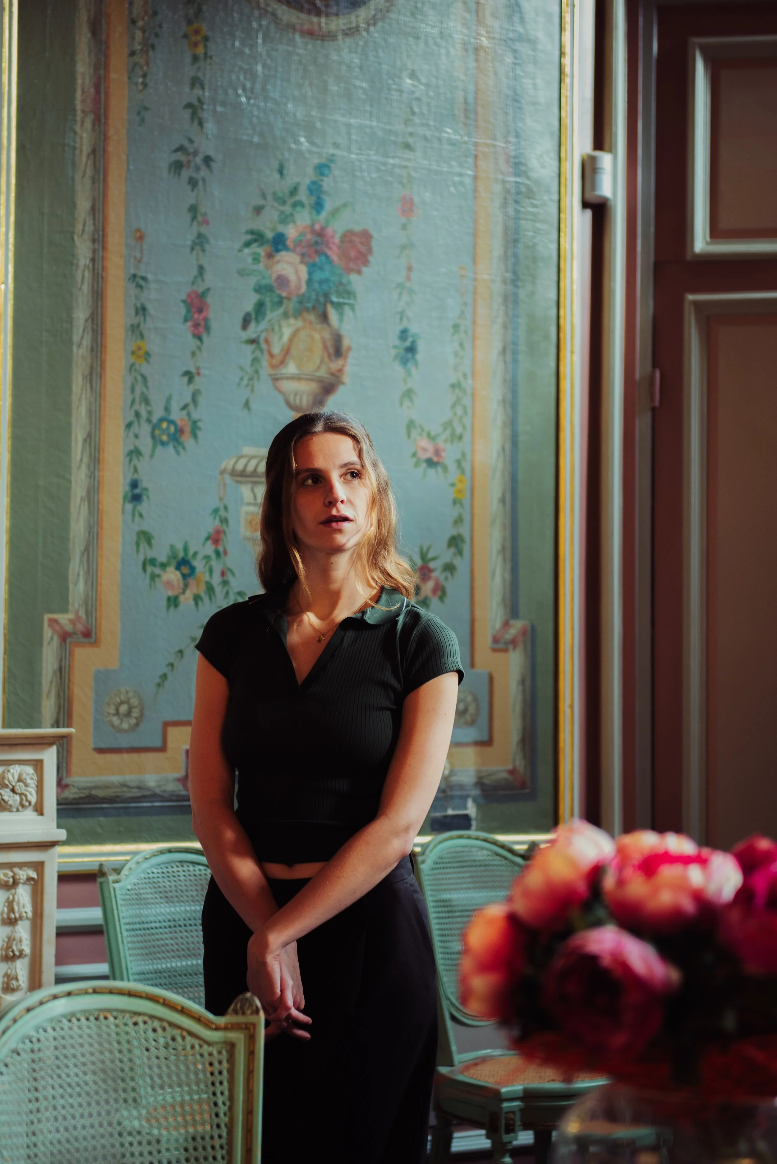 Een jonge vrouw met bruin haar staat in een elegant interieur met opvallende muurdecoratie en bloemen op tafel.