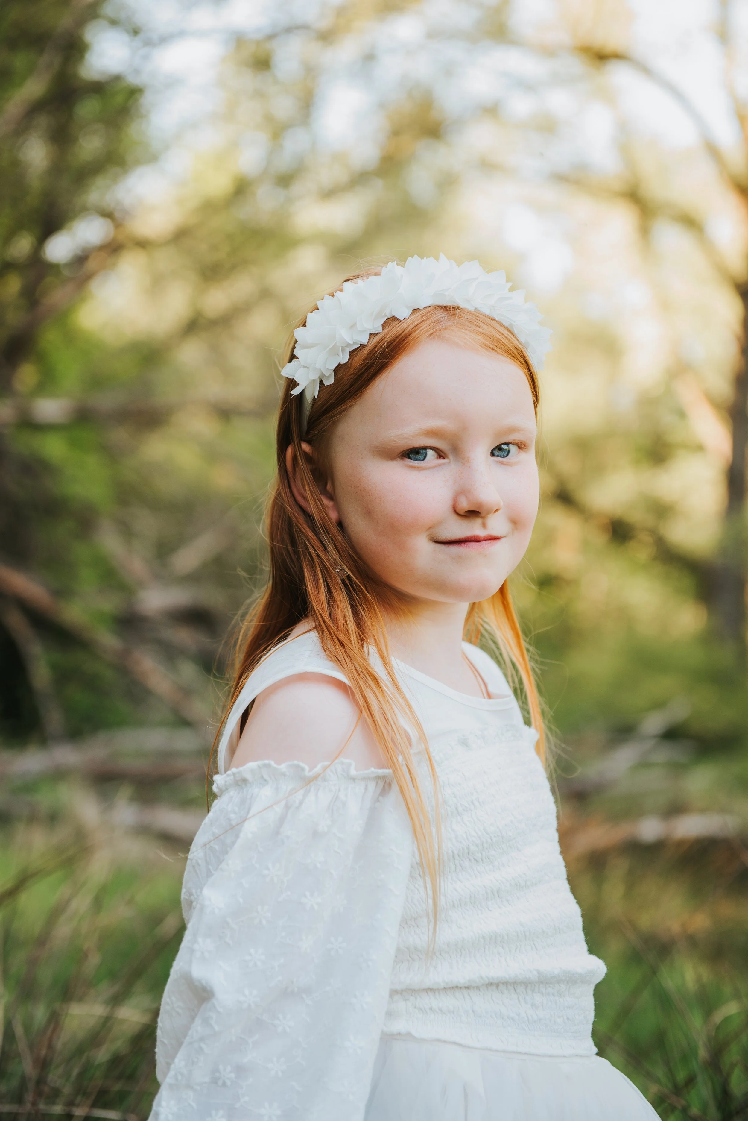 Een jonge roodharige vrouw in een witte jurk met een bloemenkroon in een bos, met een lichte en groene achtergrond.