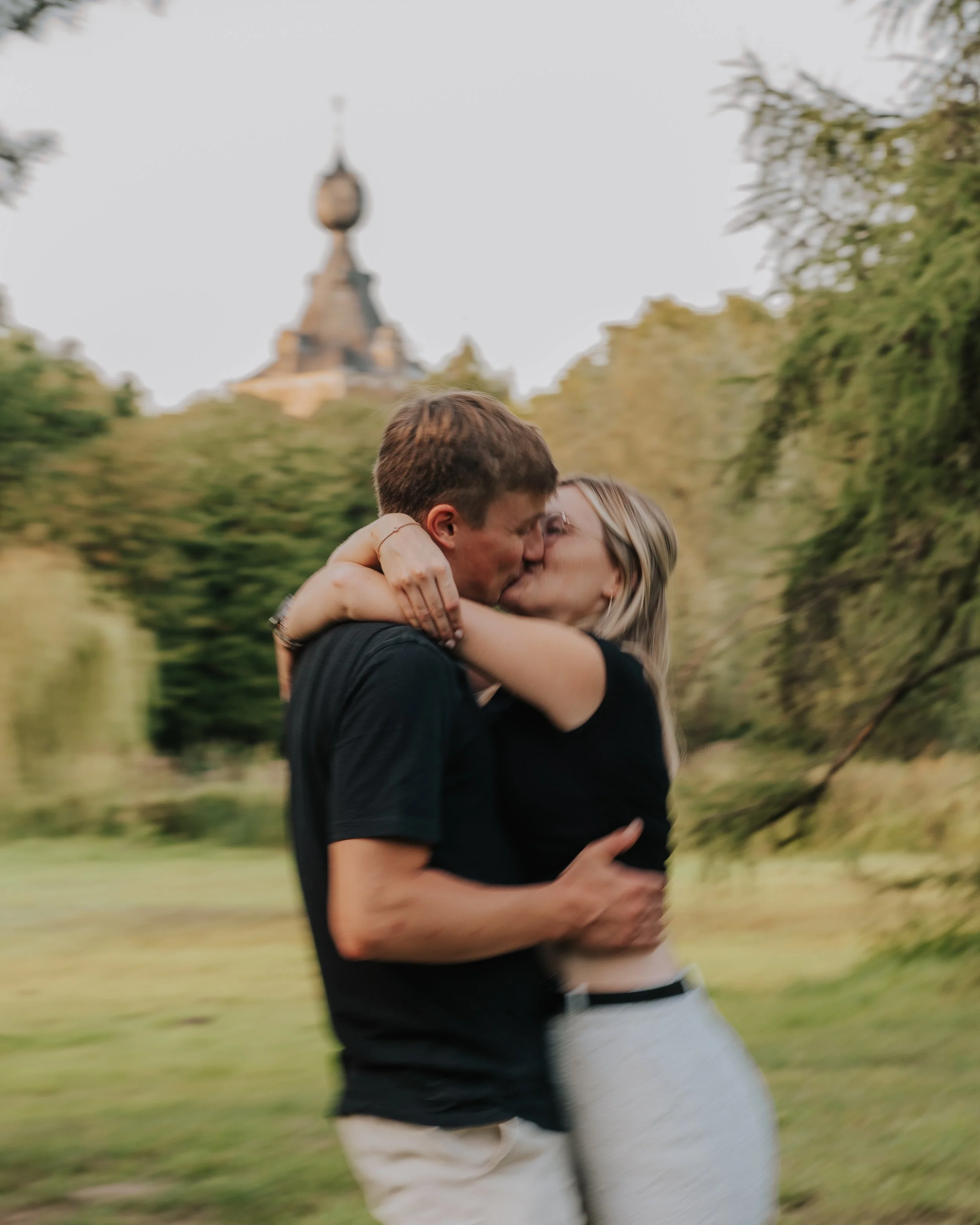 Een koppel kust elkaar terwijl ze knuffelen in een park met een historische toren op de achtergrond.