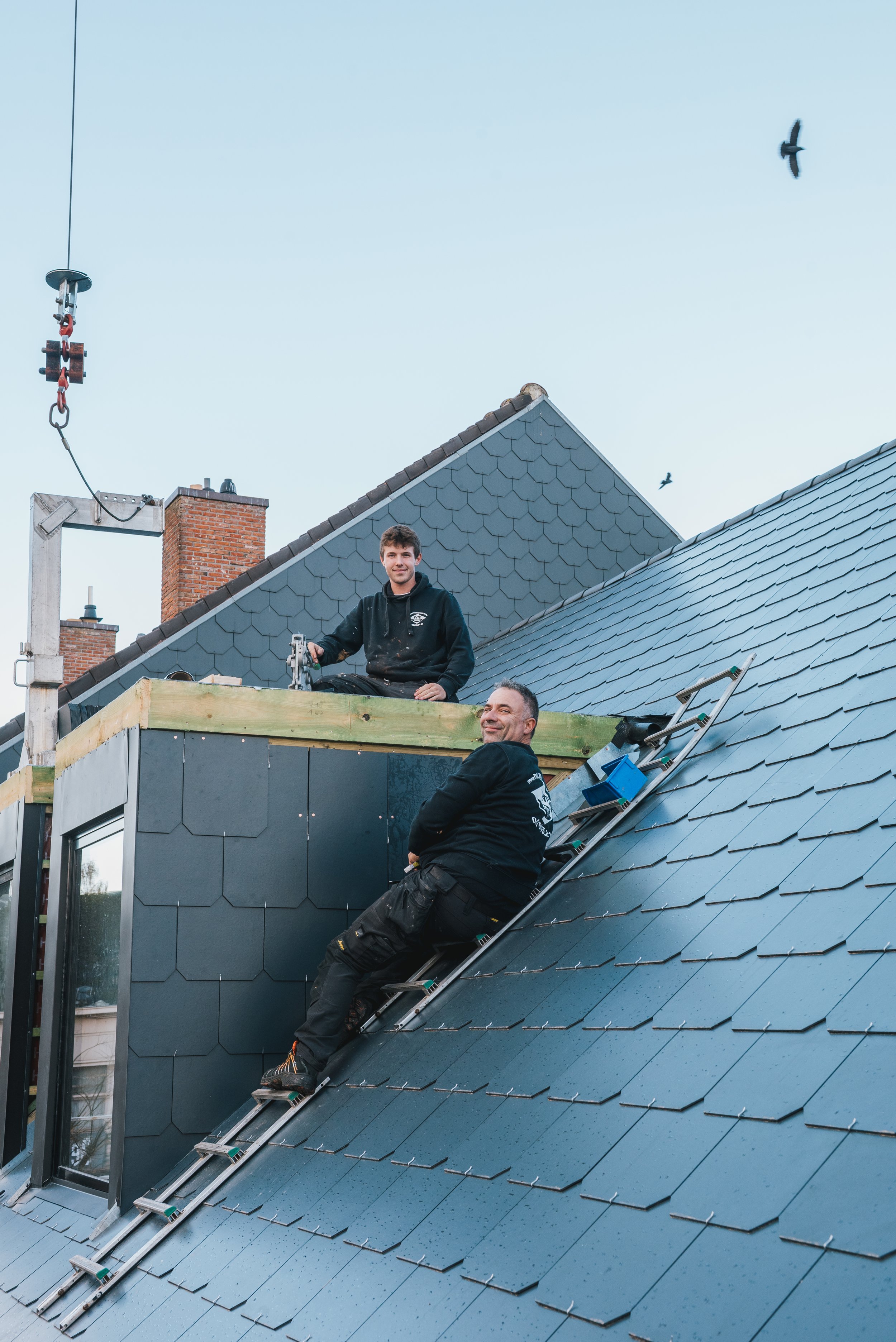Twee mannen werken op het dak van een huis, een staat op een ladder en de ander zit op het dak. Het dak heeft blauwe dakpannen en er is een grote raampartij.