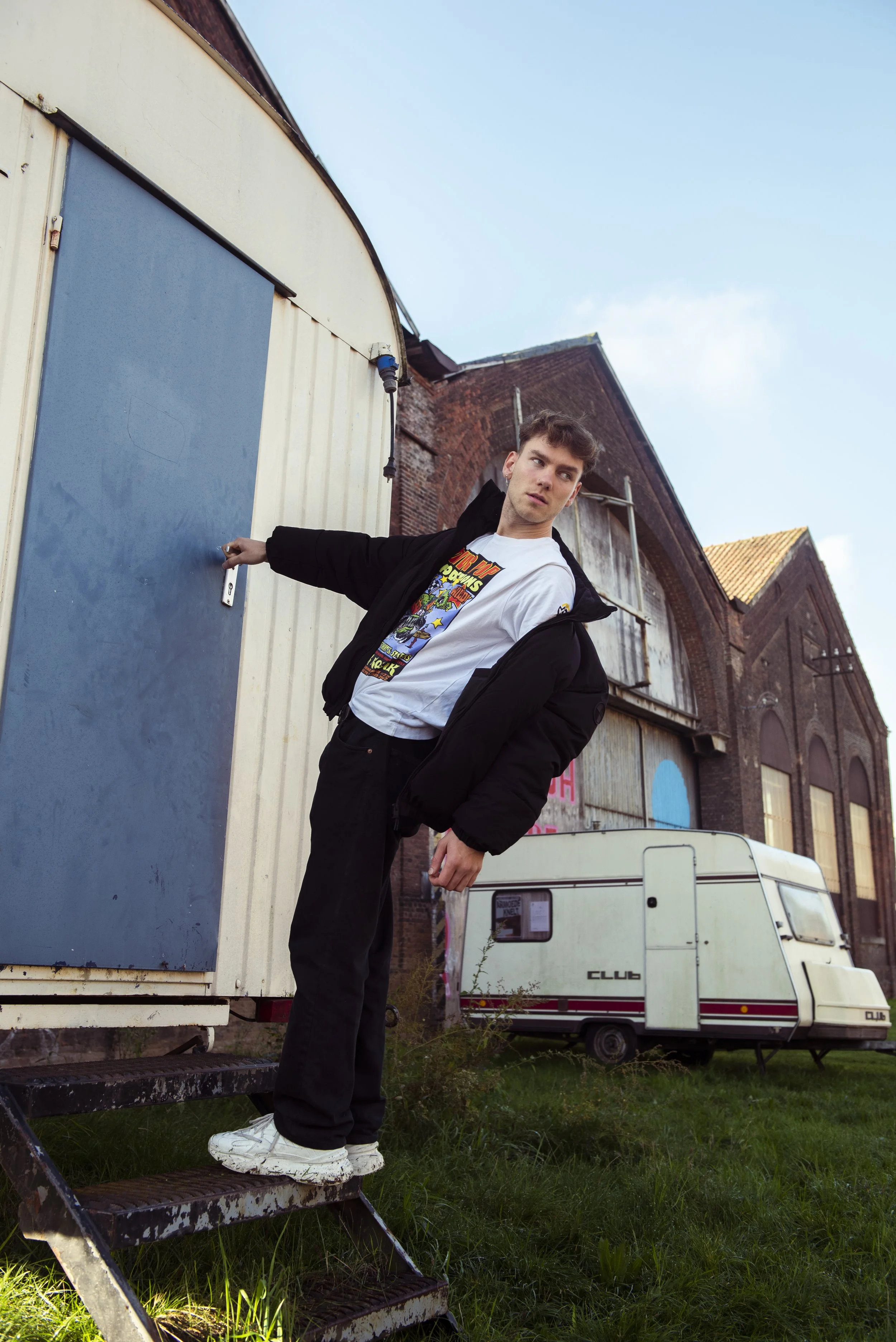 Jongeman staat op roestige metalen trap bij een blauwe deur, draagt een graphic t-shirt en een zwarte jas, met een caravan en oude bakstenen gebouwen op de achtergrond, onder een blauwe lucht.