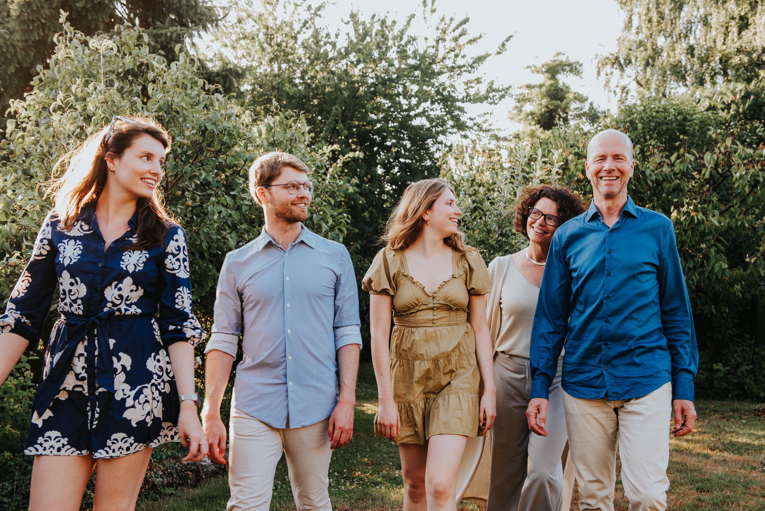 Vijf mensen wandelen samen in een tuin, lachend en genietend van de zon, omgeven door groene bomen en struiken.