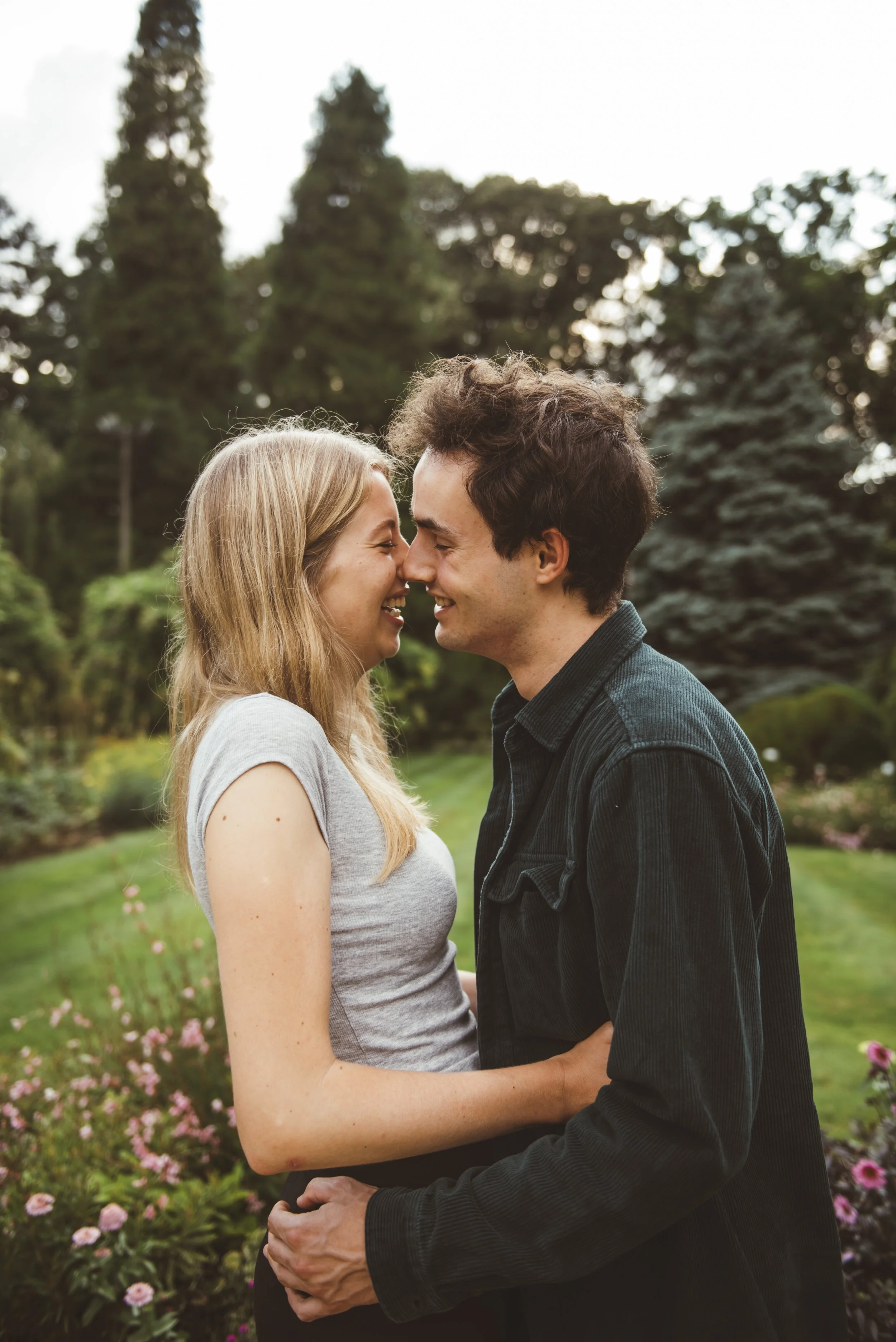 Een stel lacht naar elkaar met hun voorhoofden tegen elkaar, in een tuin met bloemen en bomen.
