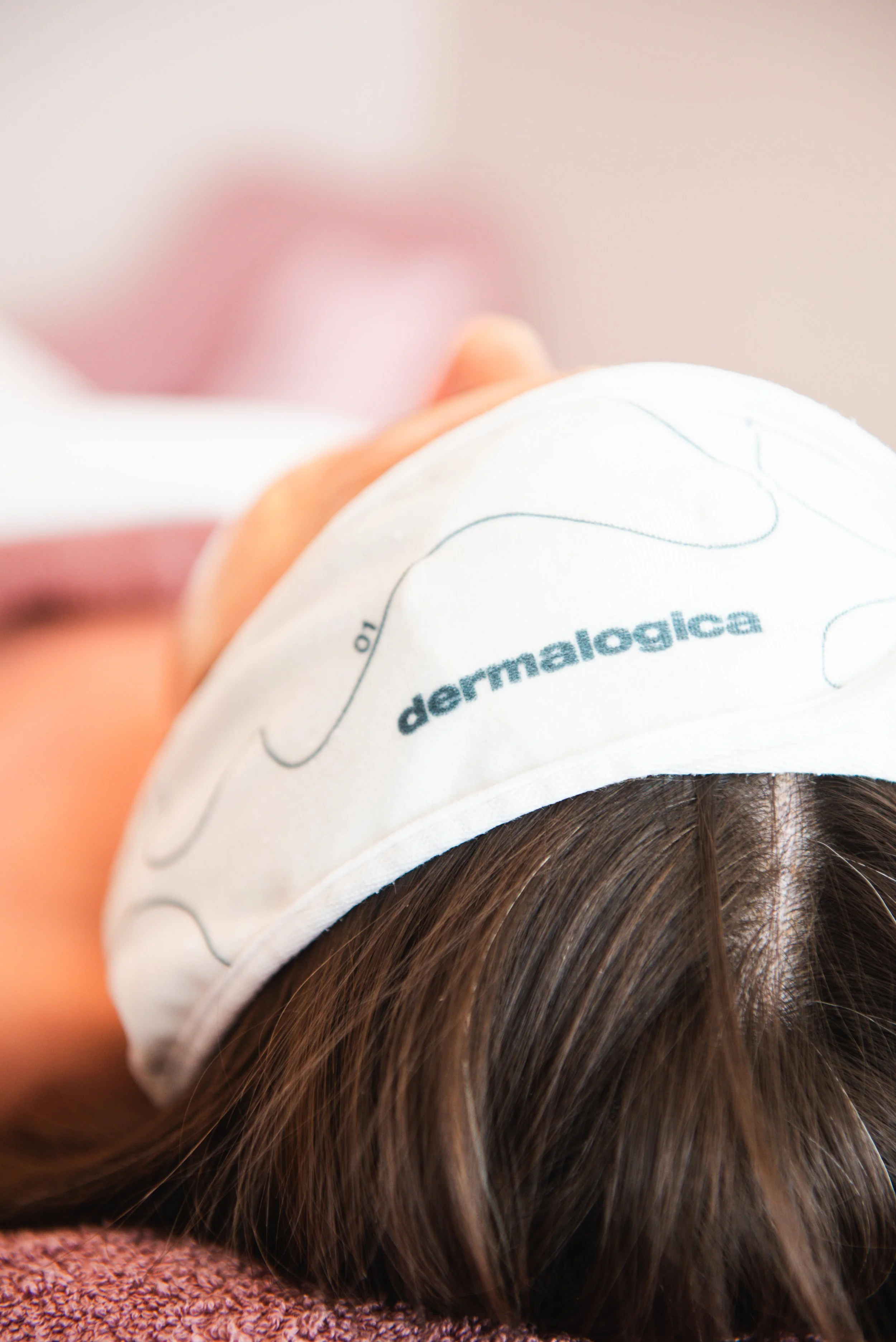 Close-up van een persoon met bruin haar, liggend op een tafel, met een wit gezichtsmasker dat 'dermalogica' heet.