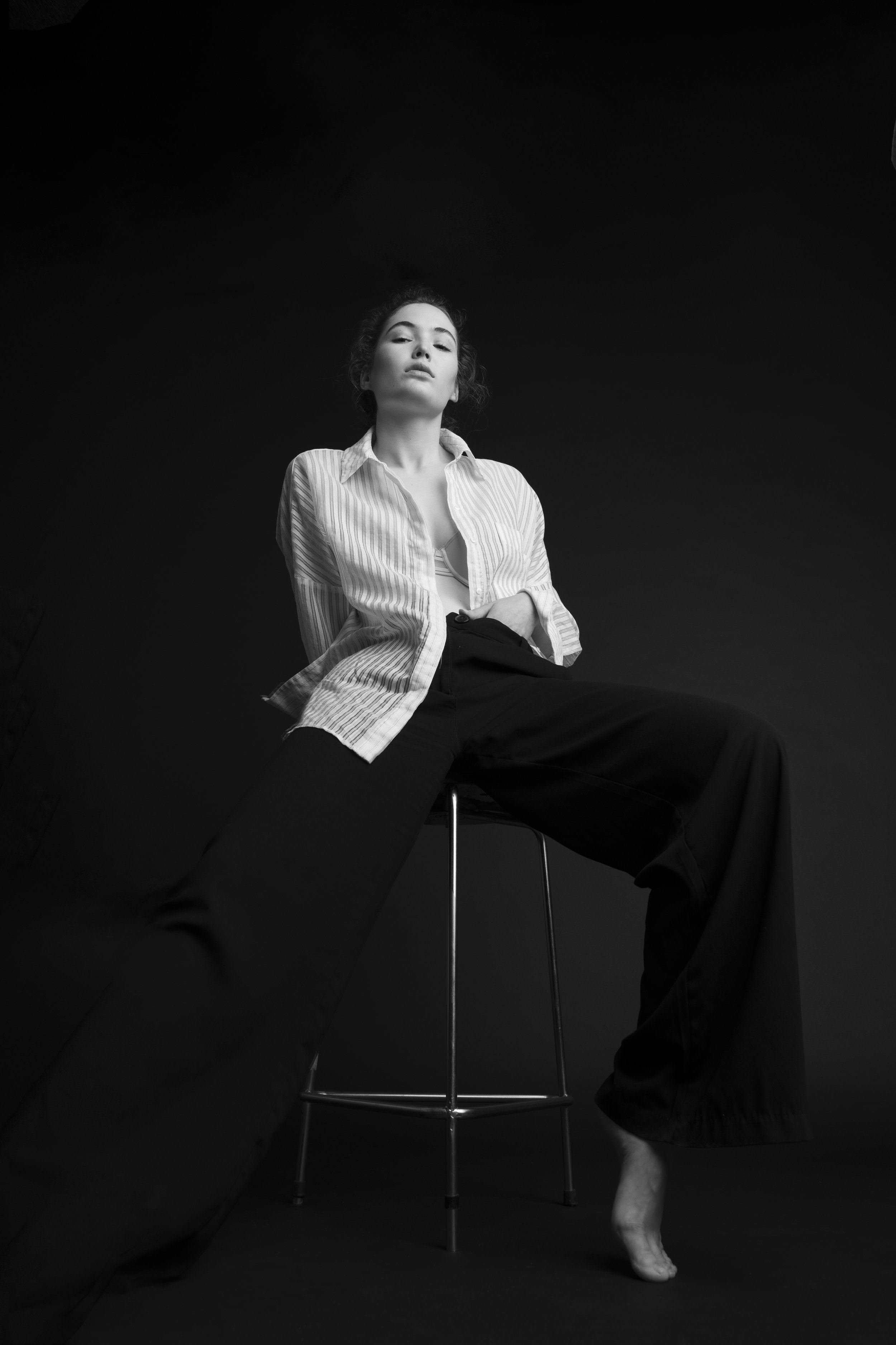 Zwart-wit foto van een vrouw zittend op een hoge kruk, met haar benen gespreid, gekleed in een blouse en wijde broek, met een serieuze blik en haar hand op haar buik