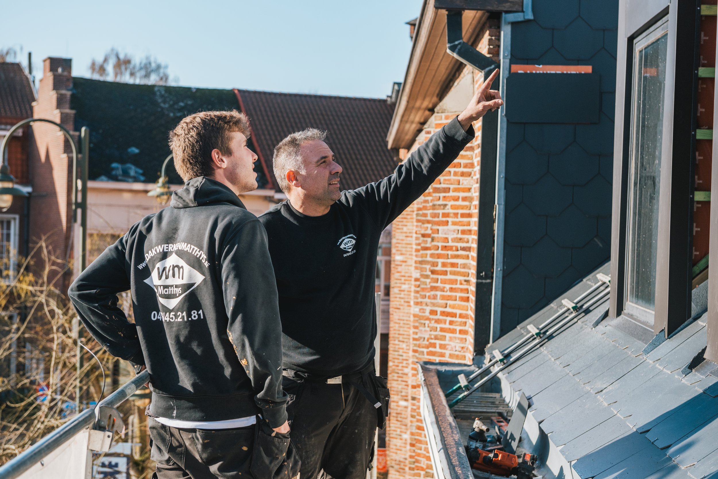 Twee bouwvakkers, één jonge en één oudere, bespreken werkzaamheden op een dak met een laptop en gereedschap.