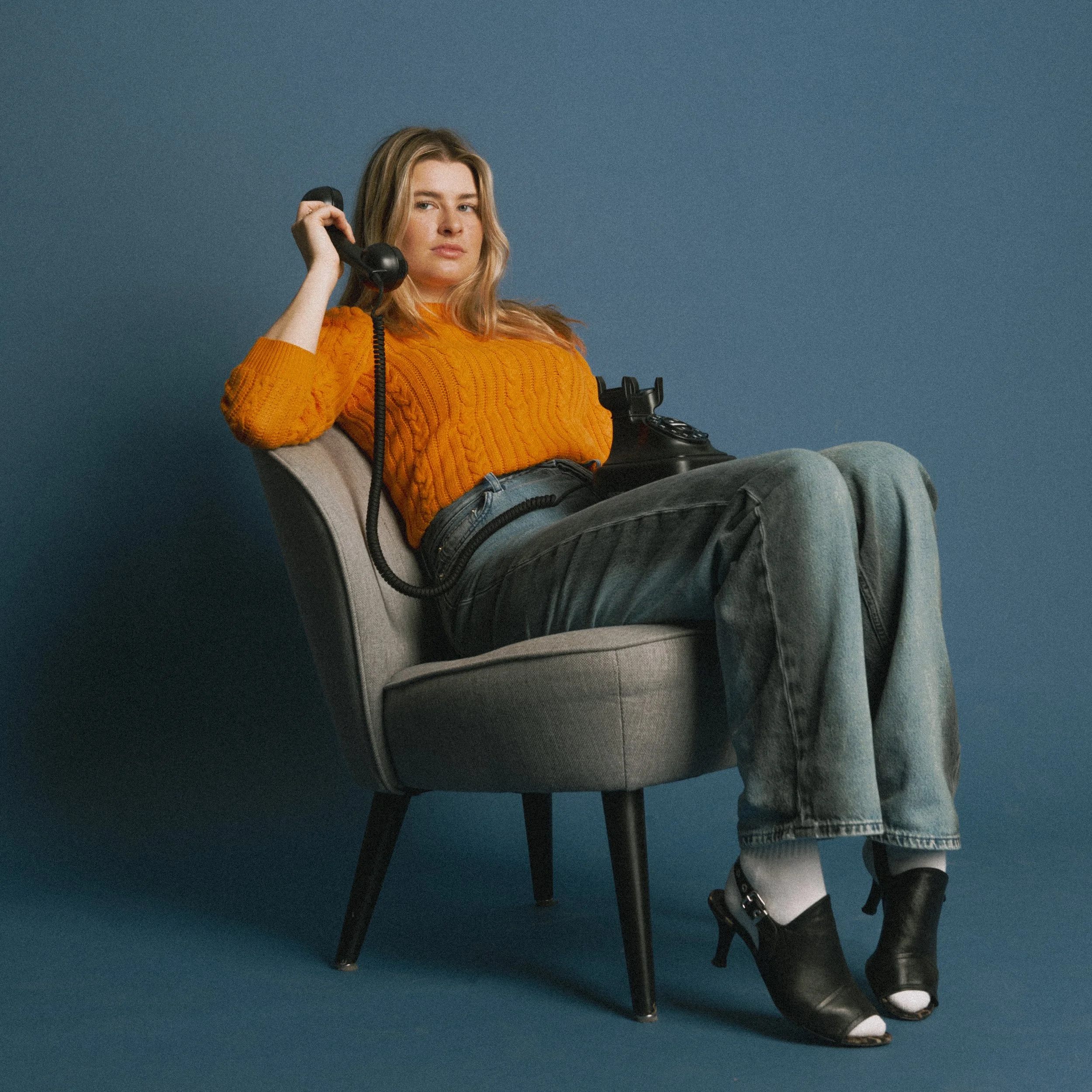 Vrouw zit op een comfortabele stoel tegen een blauwe achtergrond, draagt een oranje trui en zwarte hakken, terwijl ze een vintage zwarte telefoon vasthoudt.