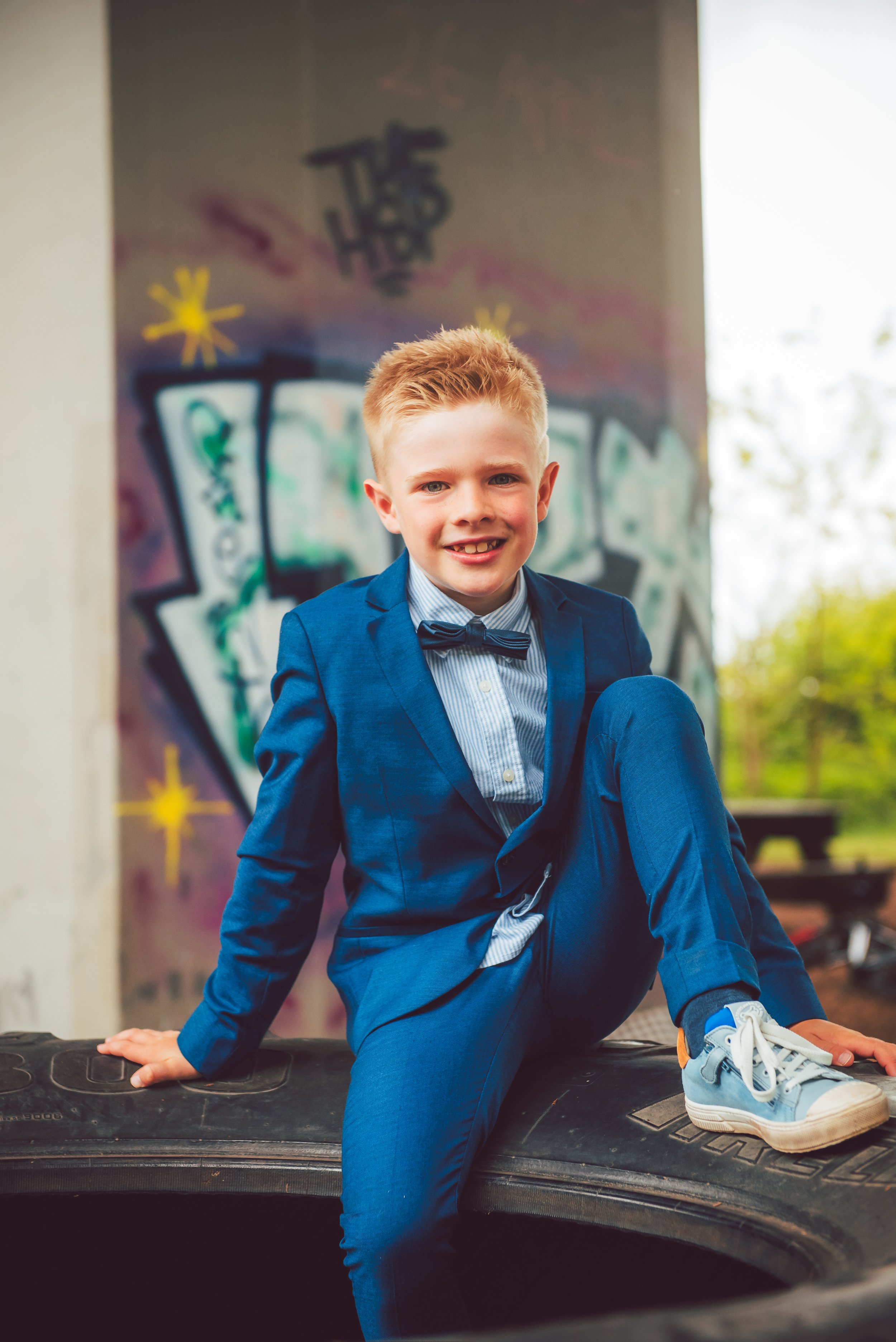 Jonge jongen in blauwe pak en stropdas zittend op een autoband bij een graffiti muurschildering in een park.