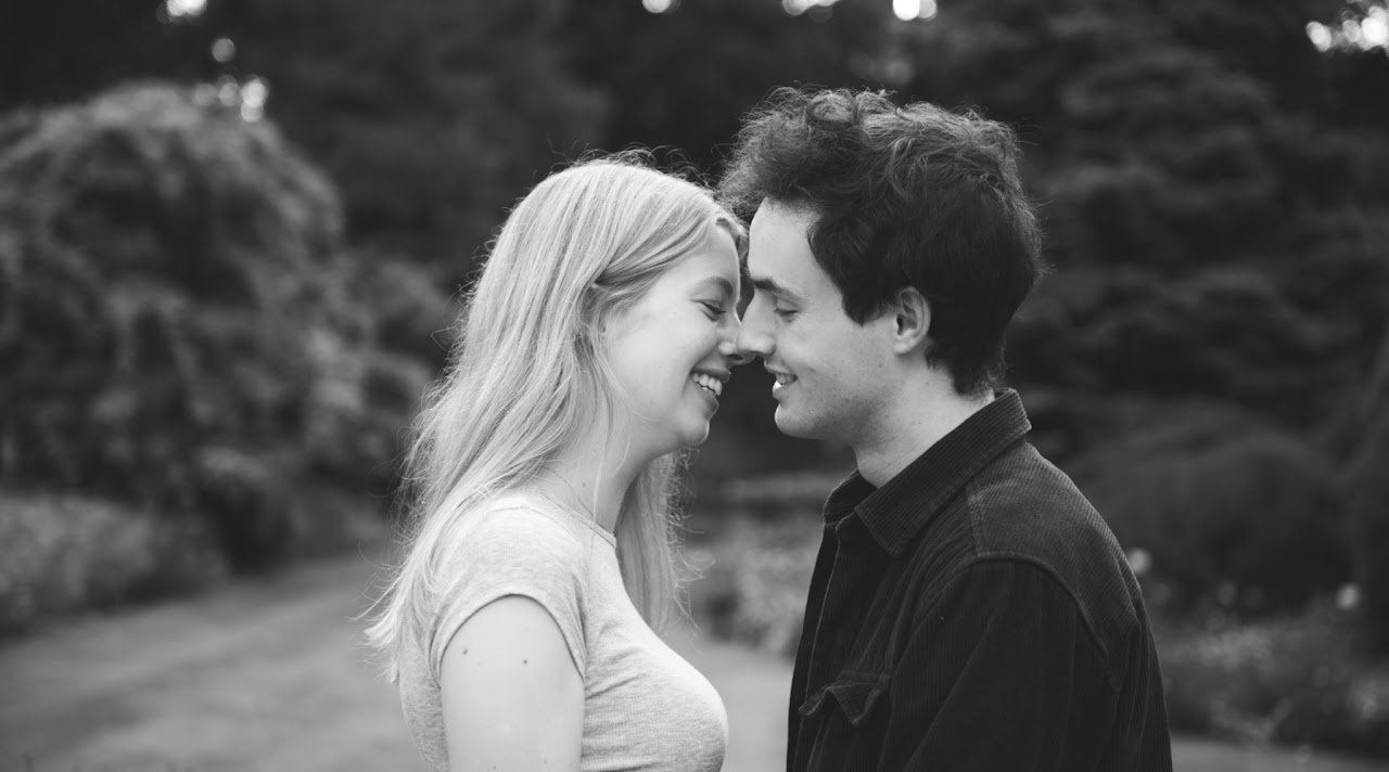 Een jonge man en een jonge vrouw staan dichtbij elkaar met hun voorhoofd tegen elkaar, glimlachend in een zwart-wit foto.