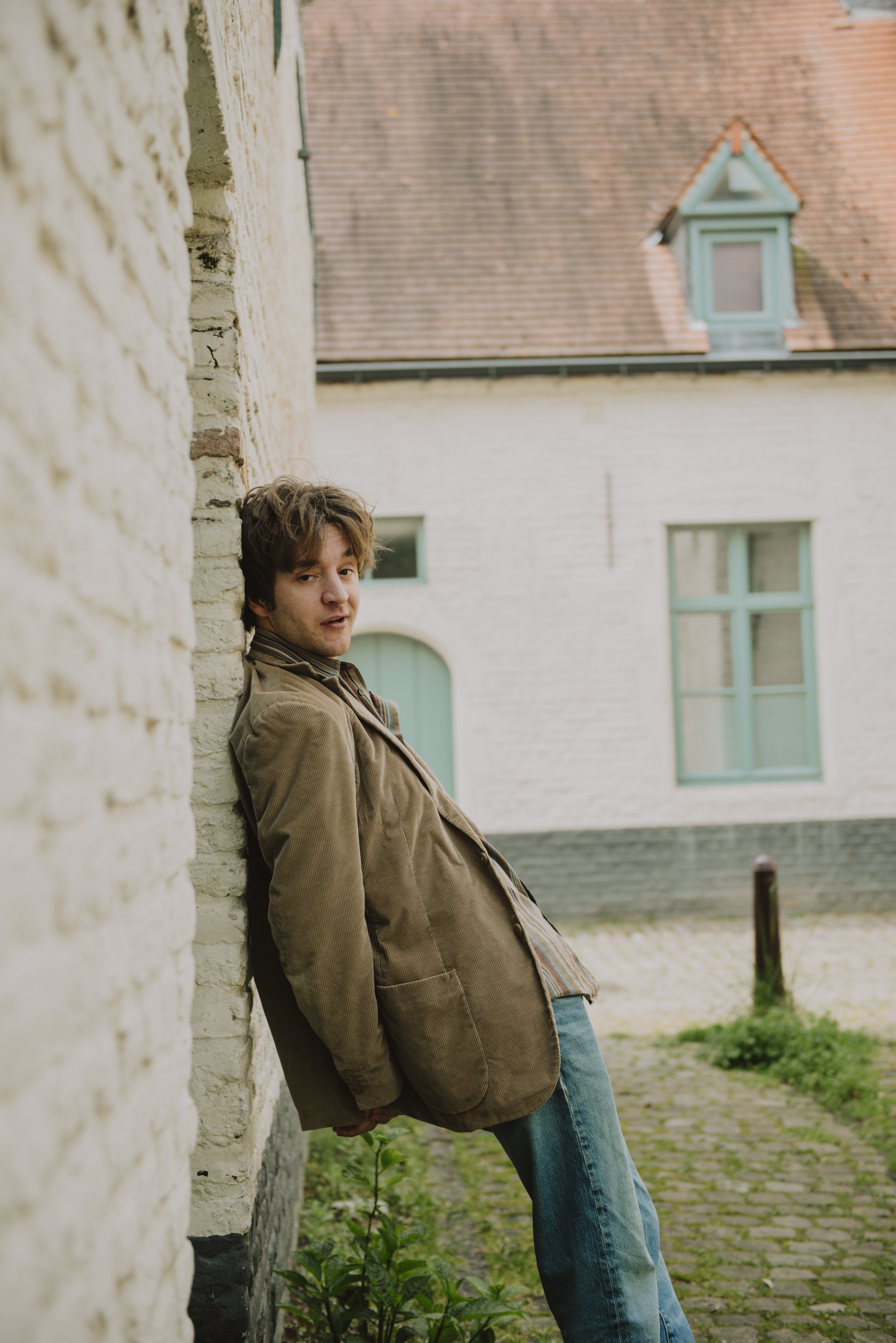Jongeman leunt tegen een muur buiten, draagt een bruin jasje en blauwe jeans, met een huis met groene ramen op de achtergrond.