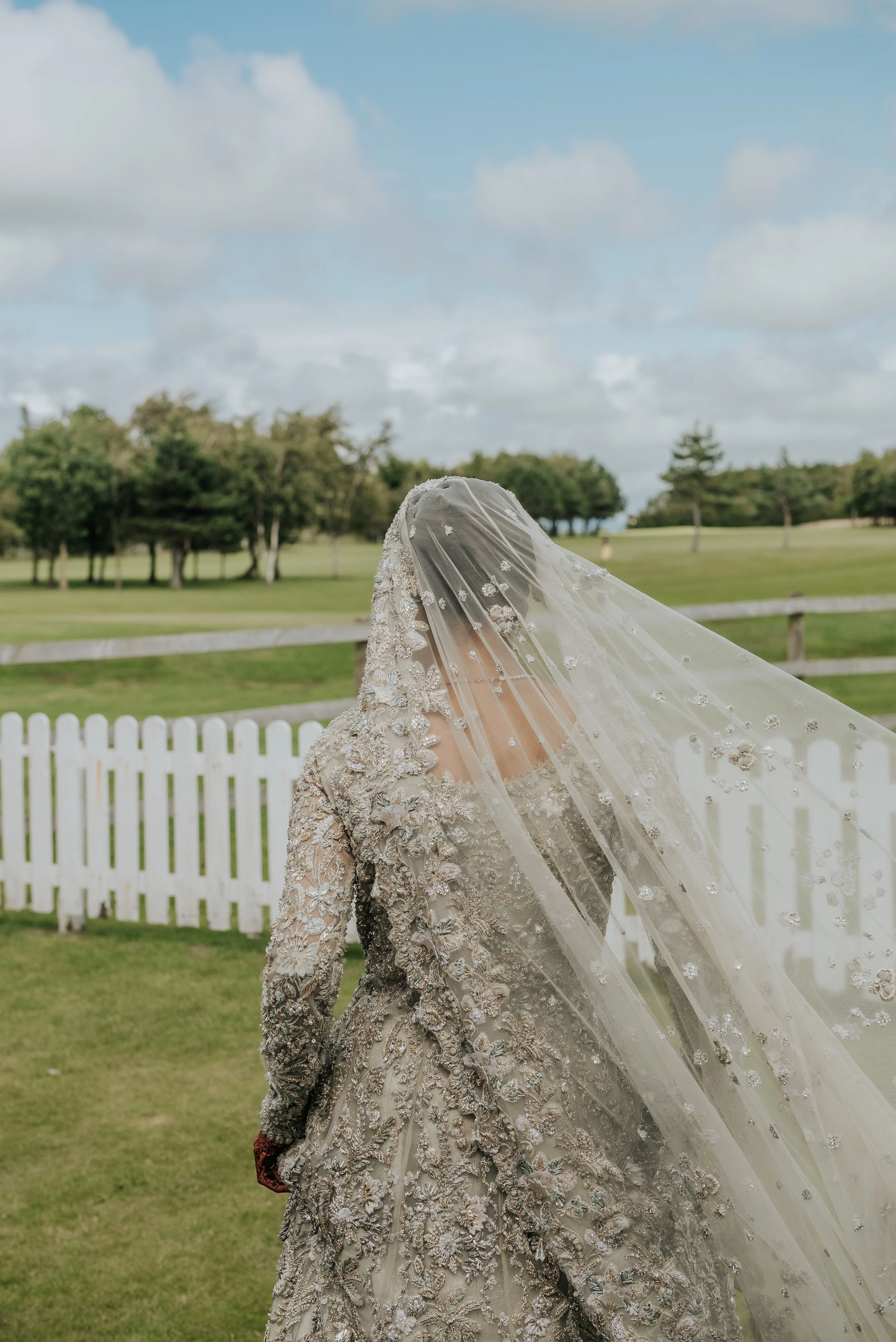 Een bruid in een gedetailleerde witte trouwjurk met kanten en pailletten, met een sluier die haar gezicht bedekt, buiten met groene bomen en een witte schutting op de achtergrond.