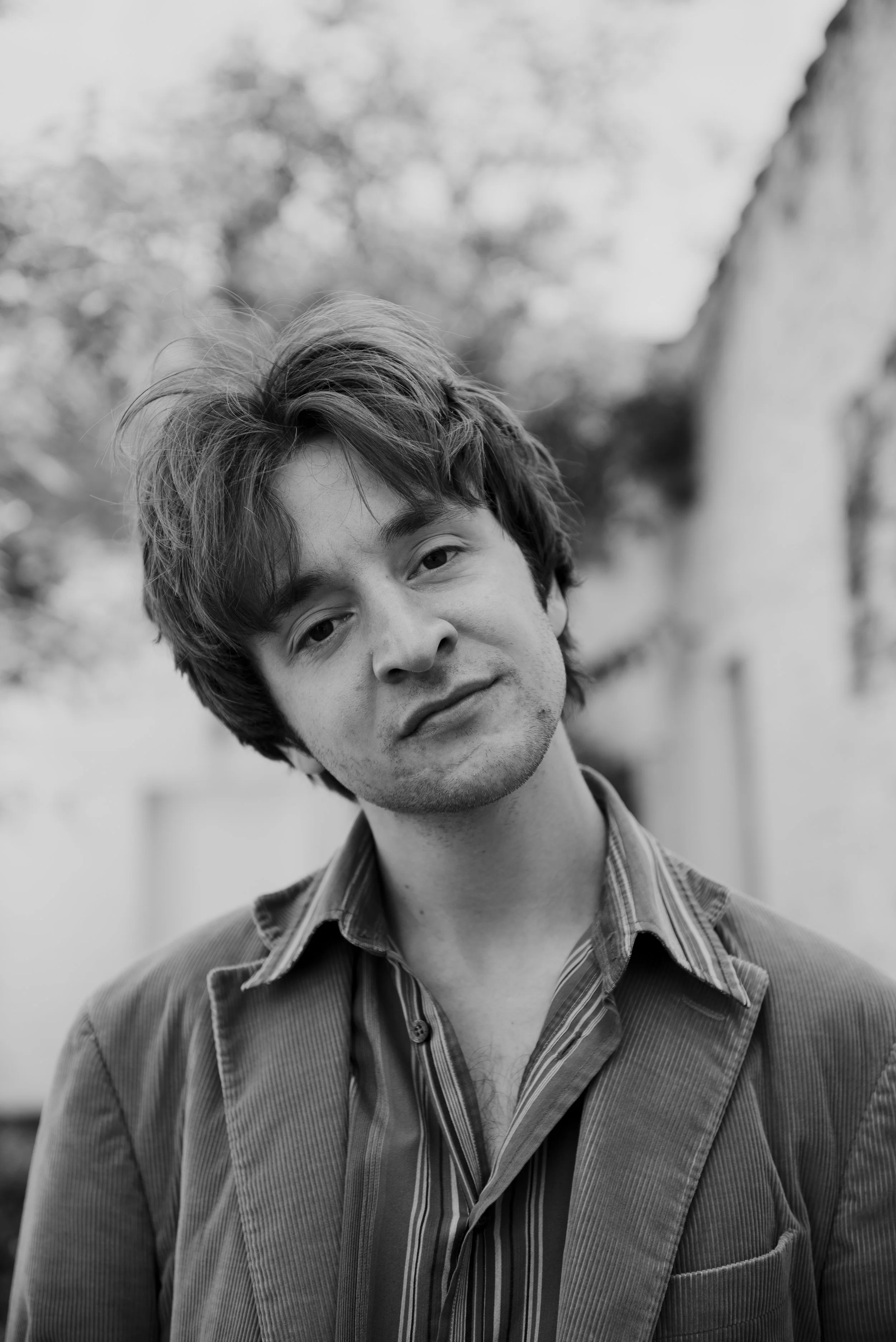 Zwart-wit foto van een jonge man met warrig haar, gekleed in een blazer en een gestreept overhemd, staand buiten met bomen en een muur op de achtergrond.