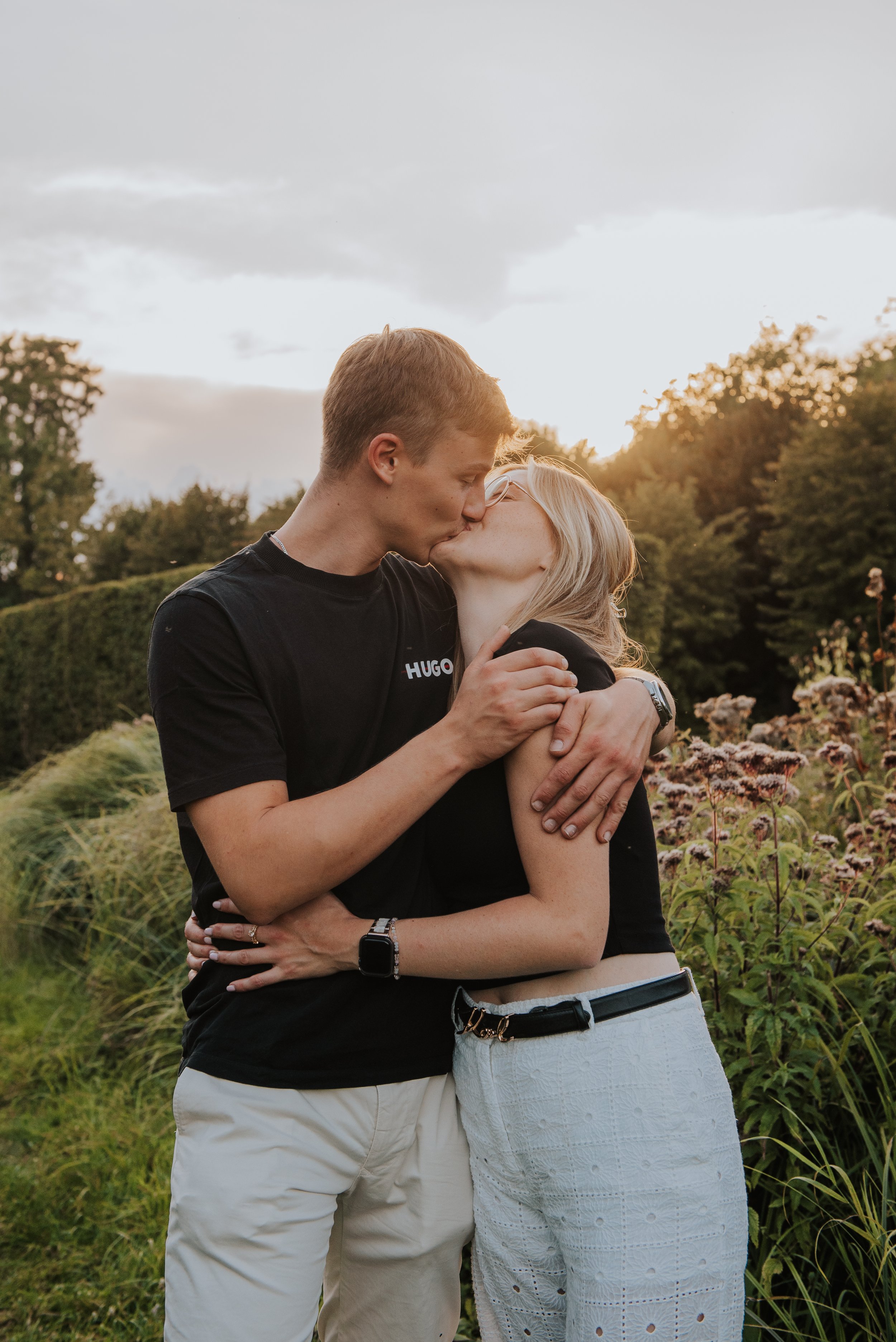 Een paar kust elkaar in een park tijdens zonsondergang, omgeven door groene planten en bomen.