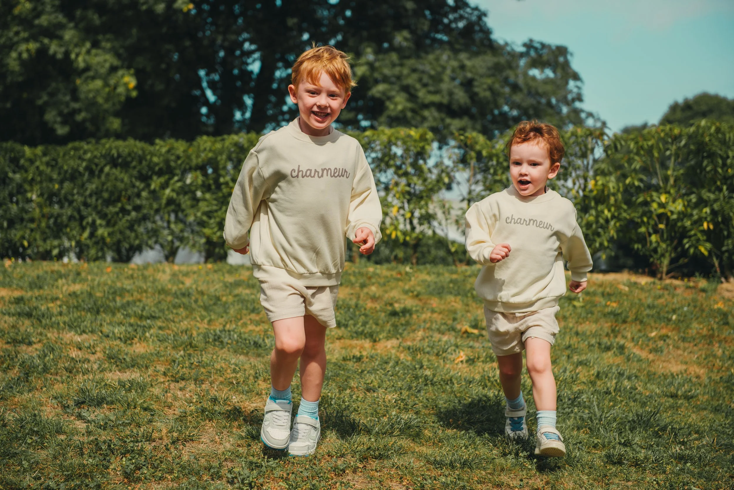 Twee jonge jongens rennen lachend over een grasveld, beide dragen roomkleurige sweatshirts en shorts, onder een groene boomrijke achtergrond.